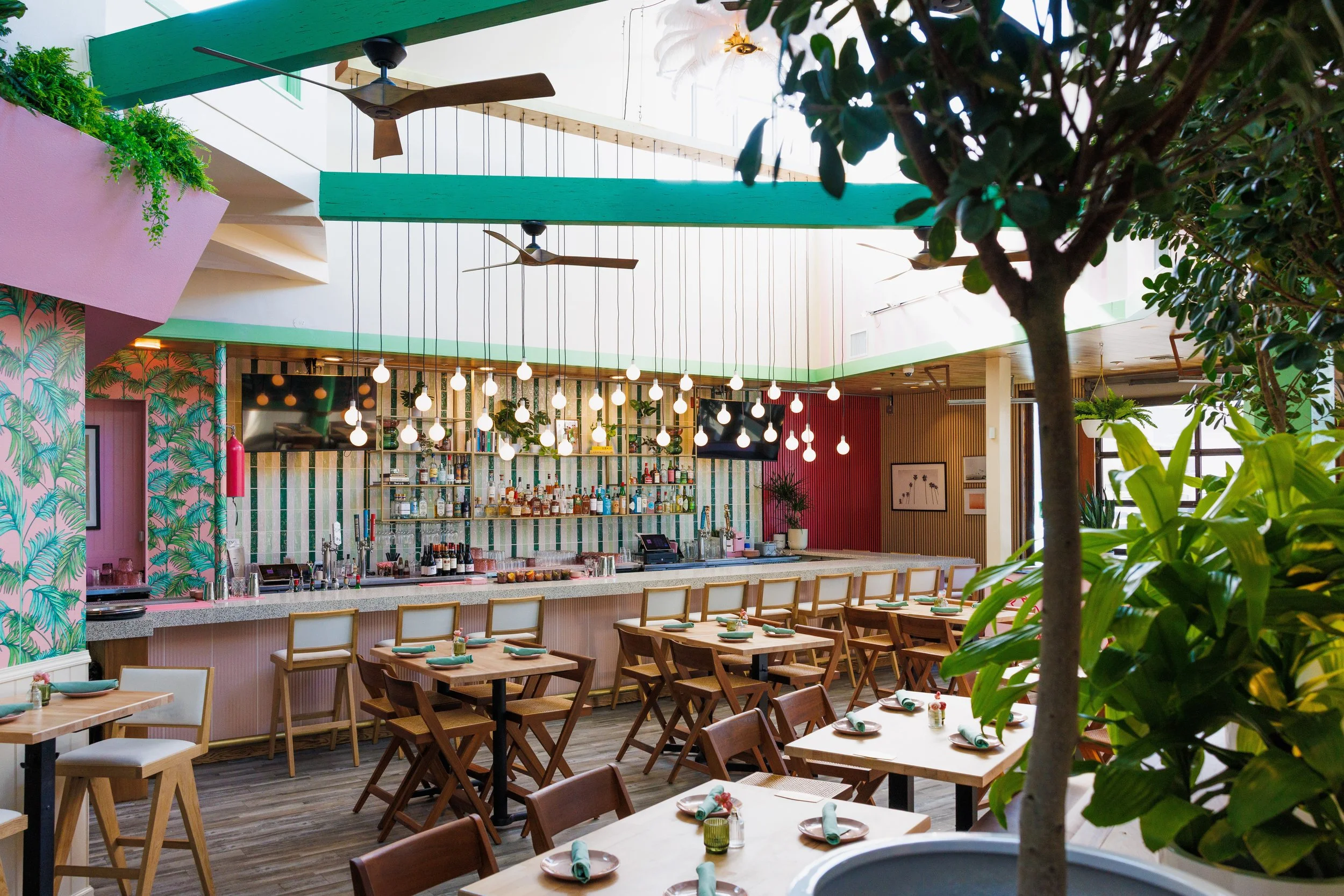 Interior of a restaurant bar with wooden chairs and tables, string lights hanging from the ceiling, a bar counter with bottles, tropical-themed wallpaper, and planters with greenery.