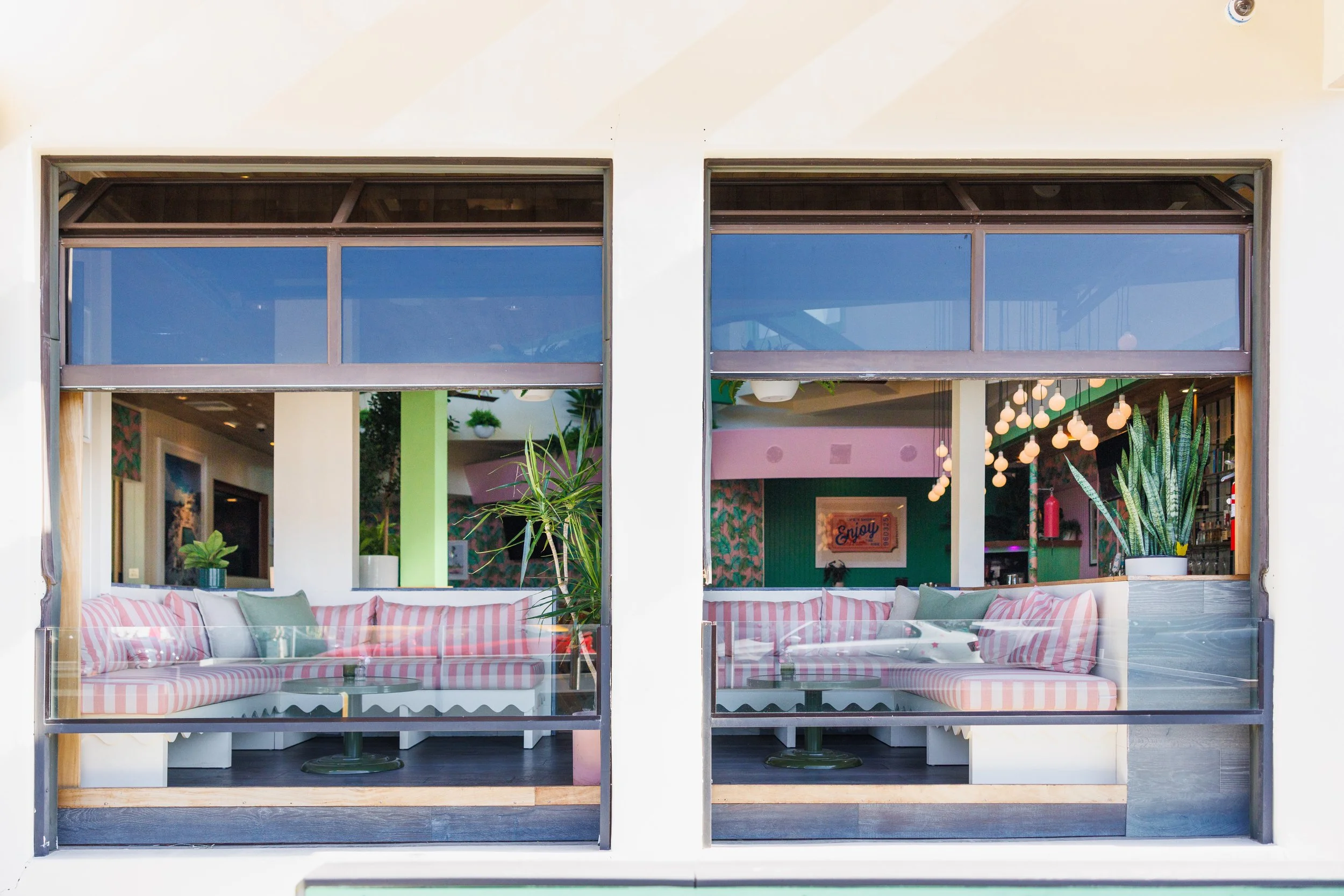 View of a stylish indoor-outdoor café with pink and white striped cushioned seating, green and pink interior walls, potted plants, and decorative hanging lights, seen through large glass windows.