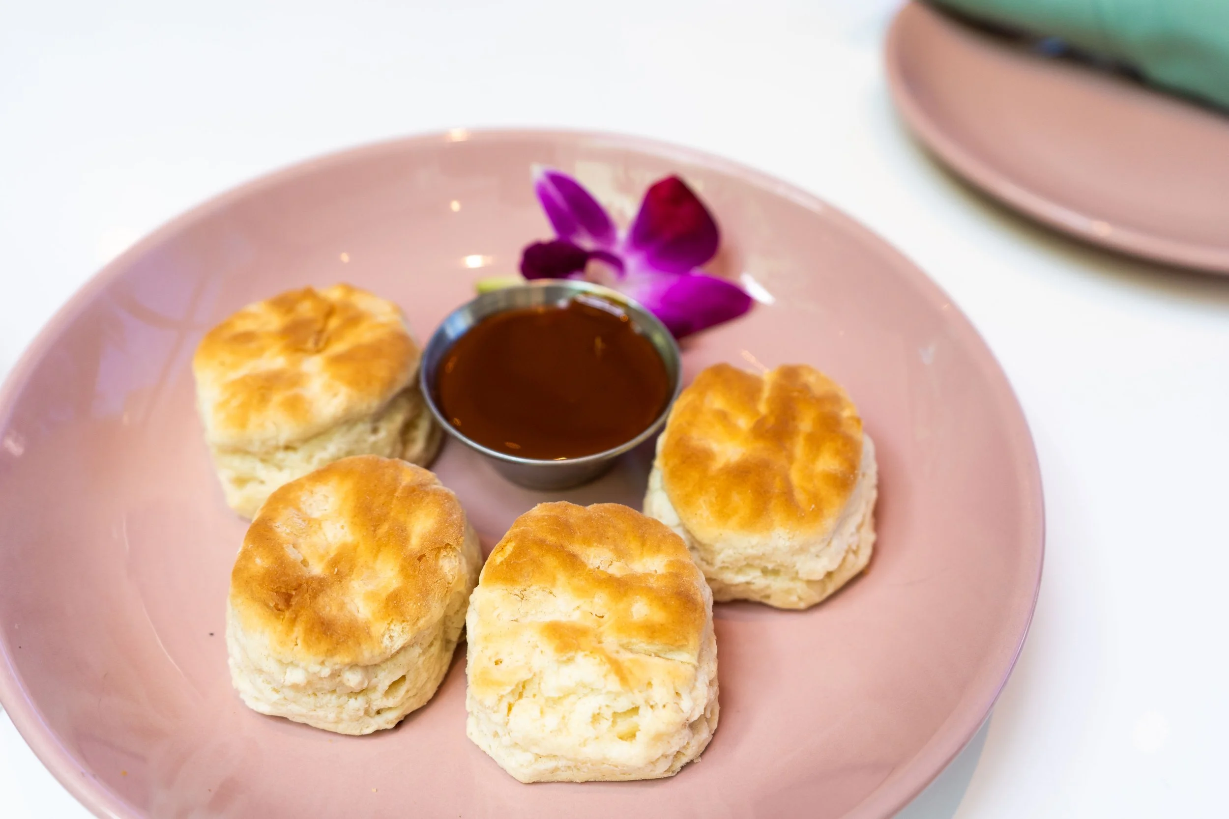 Plate with six baked scones, a small container of jam, and a purple flower, on a white surface.