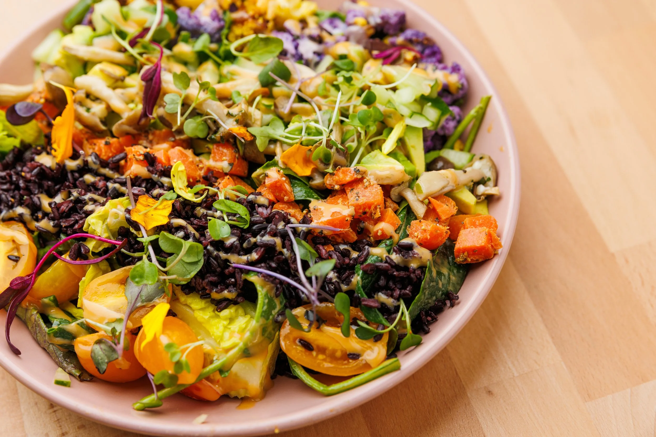 Colorful mixed vegetable salad with microgreens, black rice, cherry tomatoes, sweet potatoes, cucumbers, and other vegetables in a pink bowl on a wooden surface.