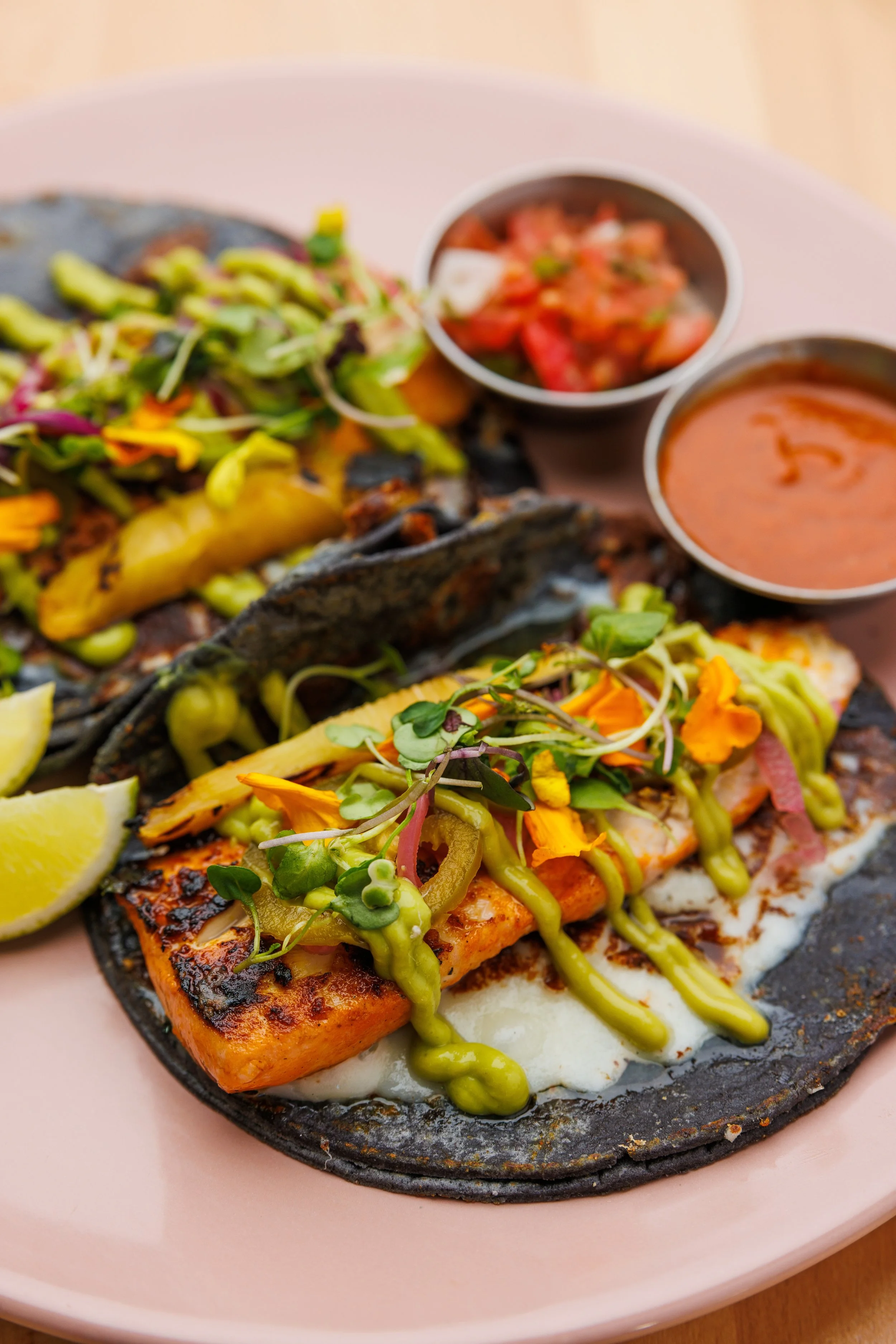 Grilled fish tacos with cheese and onions and jalapenos on a pink plate with lime wedge, topped with microgreens, served with two small bowls of salsa and sauce.