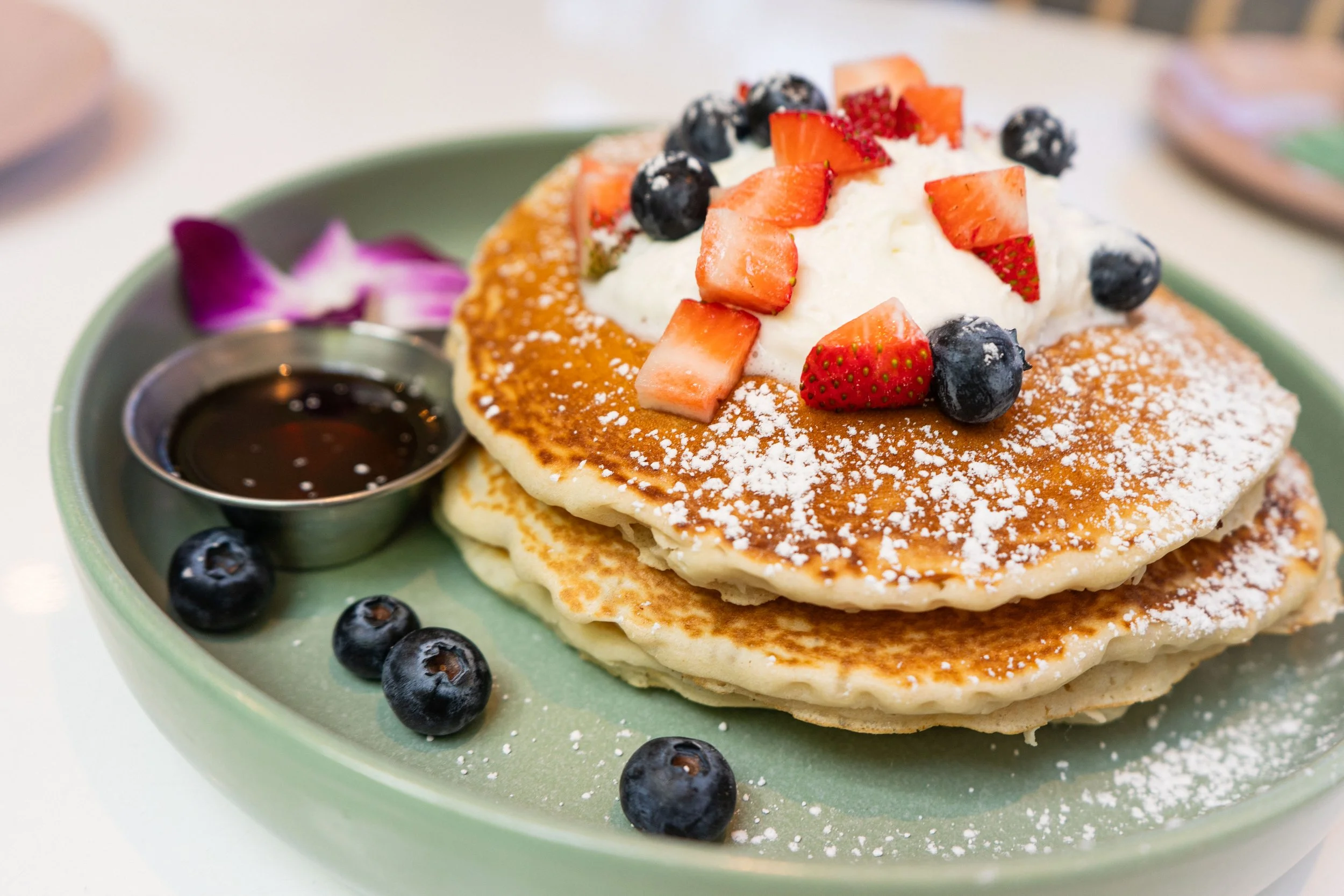 Two pancakes topped with whipped cream, sliced strawberries, and blueberries, served on a green plate with a small container of syrup and additional blueberries, with purple flower petals in the background.
