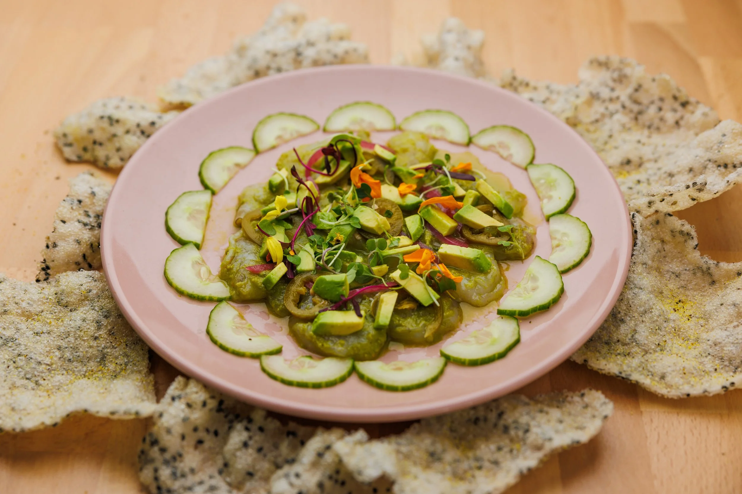 A pink plate of sliced shrimp in green salsa with chopped avocado and cucumber slices around the edge with flower leaves surrounded by rice crackers on a wooden surface.