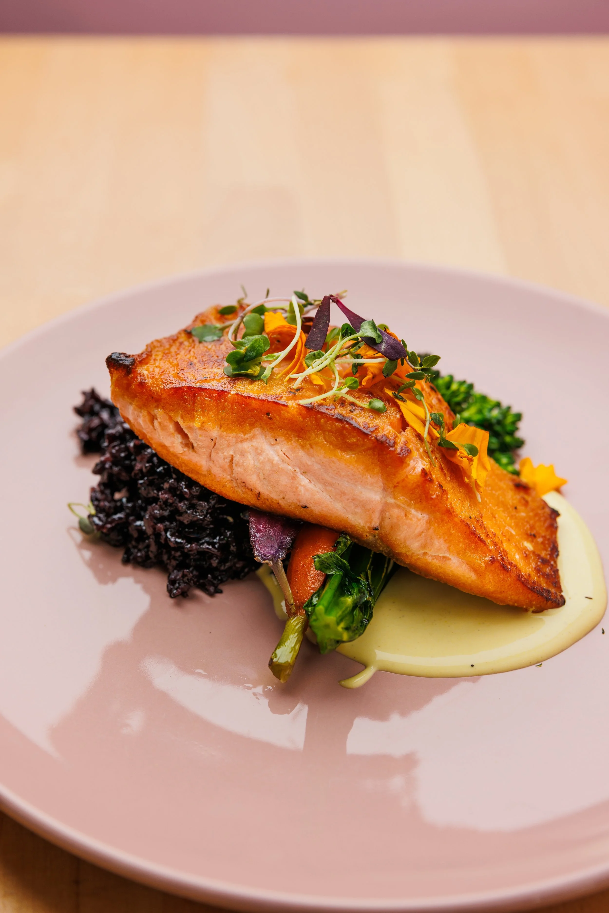 A plated dish with a piece of cooked salmon on a bed of black rice, garnished with microgreens, roasted vegetables, and a light sauce.
