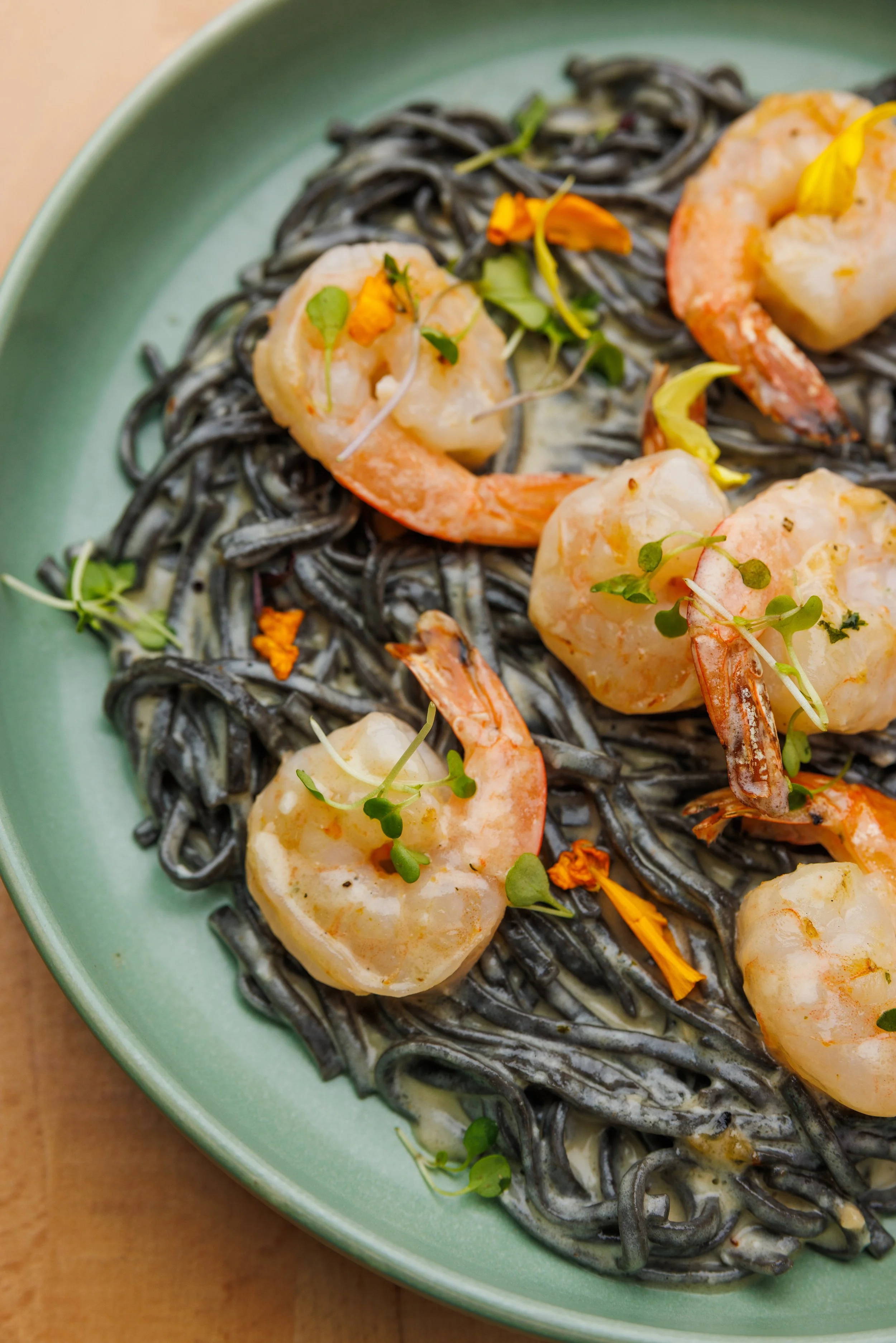 A green plate of black squid ink pasta topped with shrimp, scallops, microgreens, and edible flowers.