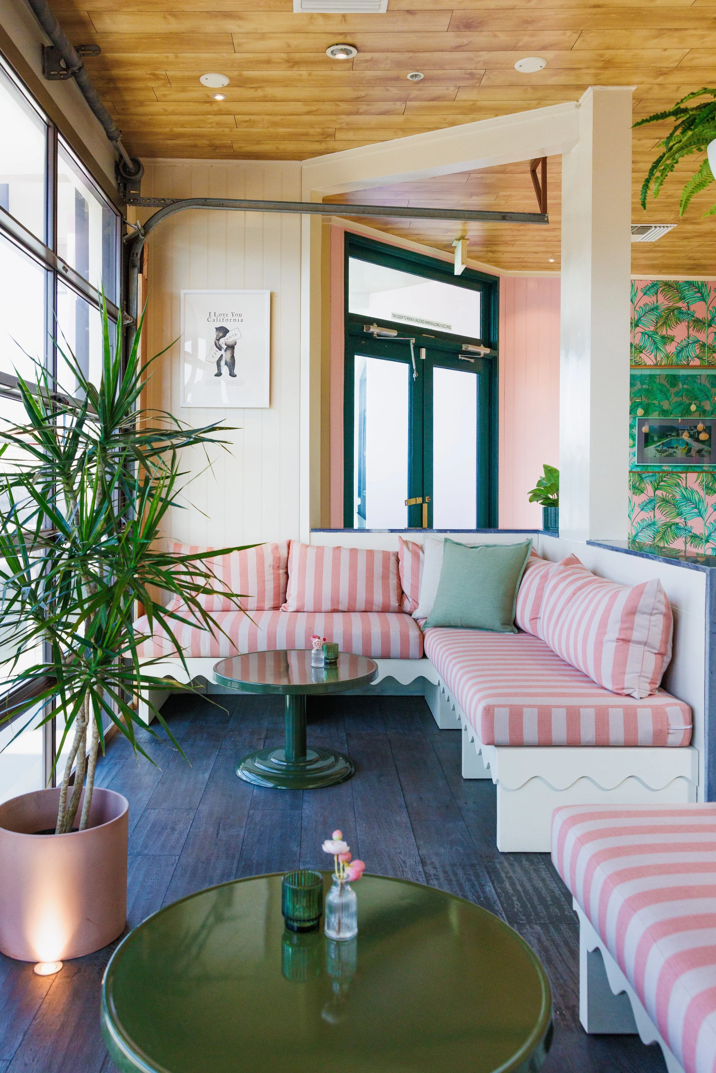 Cozy cafe corner with pink and white striped cushioned seating, green tables, potted plants, wooden ceiling, and tropical patterned wall.