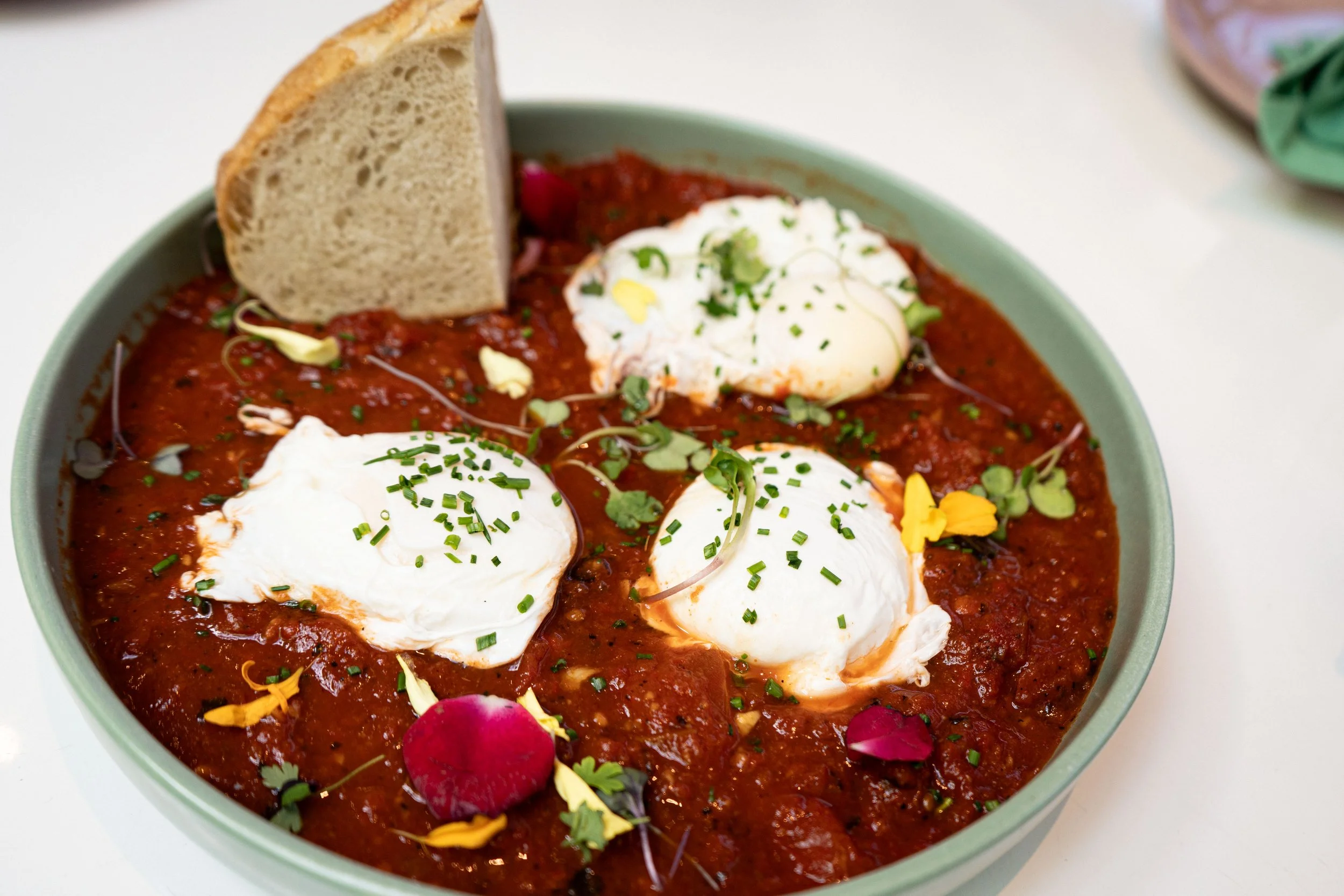 A bowl of chili topped with poached eggs, chopped chives, microgreens, and edible flower petals, with a slice of bread on the side.