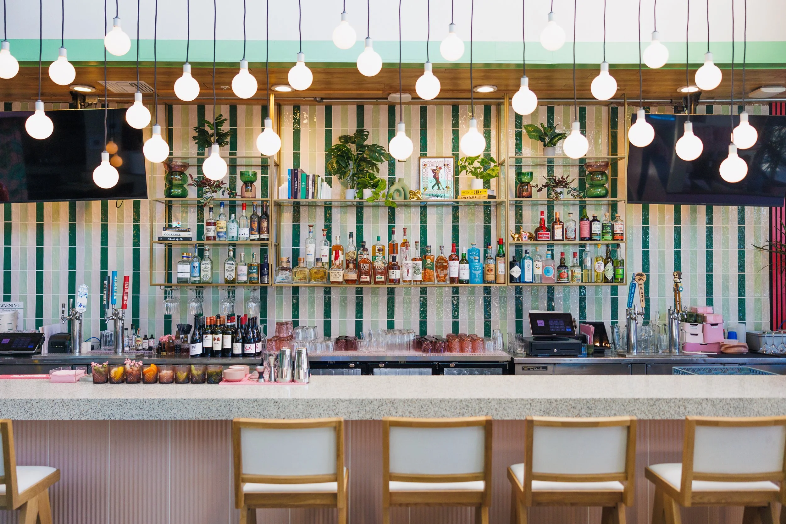 Bar with shelves of alcohol bottles, liquor, and decorative plants, with hanging pendant lights, a row of chairs, and a large flat-screen TV.