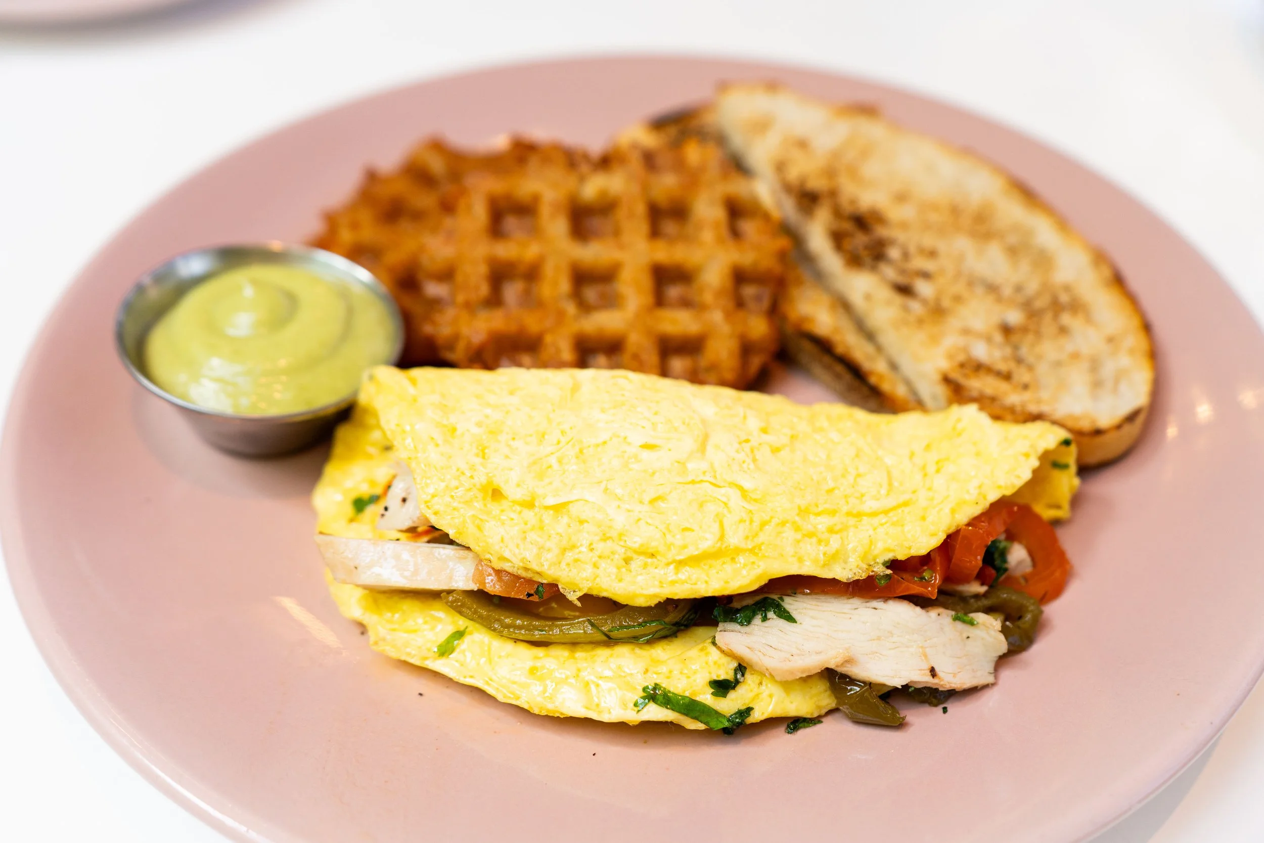 A plate of food containing a waffle, a breakfast sandwich with eggs, cheese, and vegetables, and a small cup of green sauce or dip.