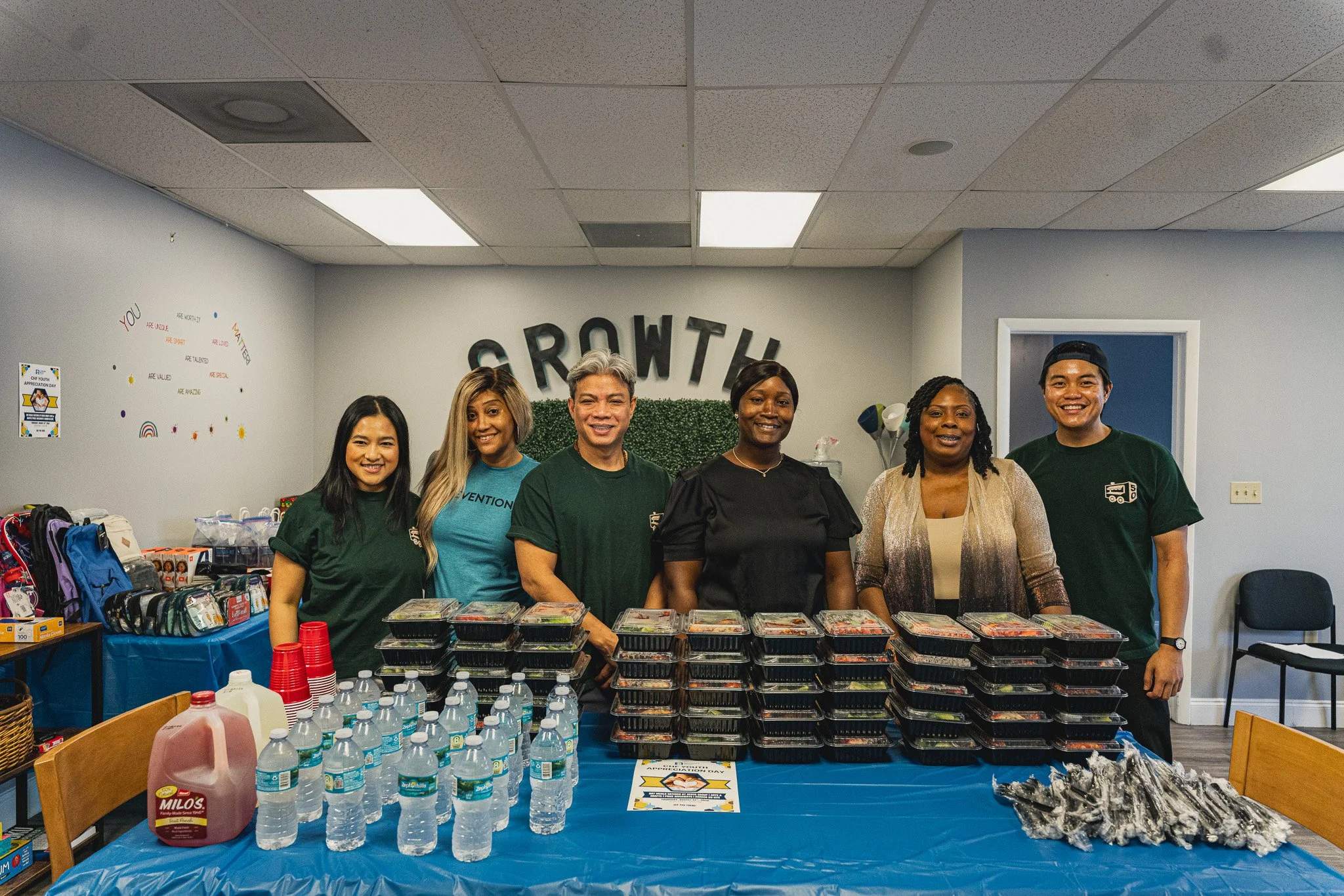 6 individuals smiling and posing for a photo in front of a table of boxed meals, water bottles and juice jugs. 