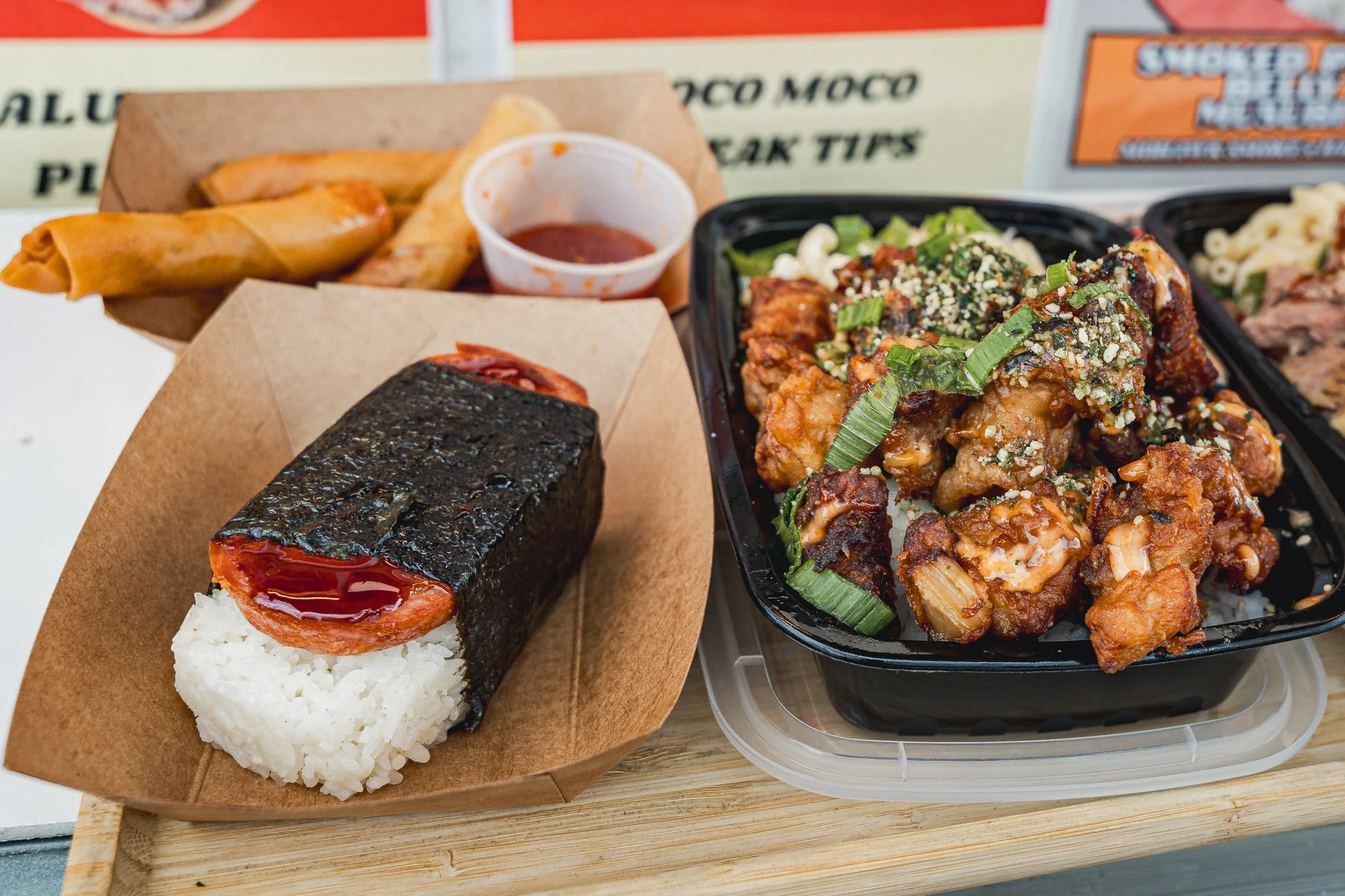Close-up of a takeout tray with rice and a sushi roll wrapped in seaweed, topped with sauce, and a side of crispy chicken with green onions and sesame seeds, along with fried chicken wings and potato chips with dipping sauce.