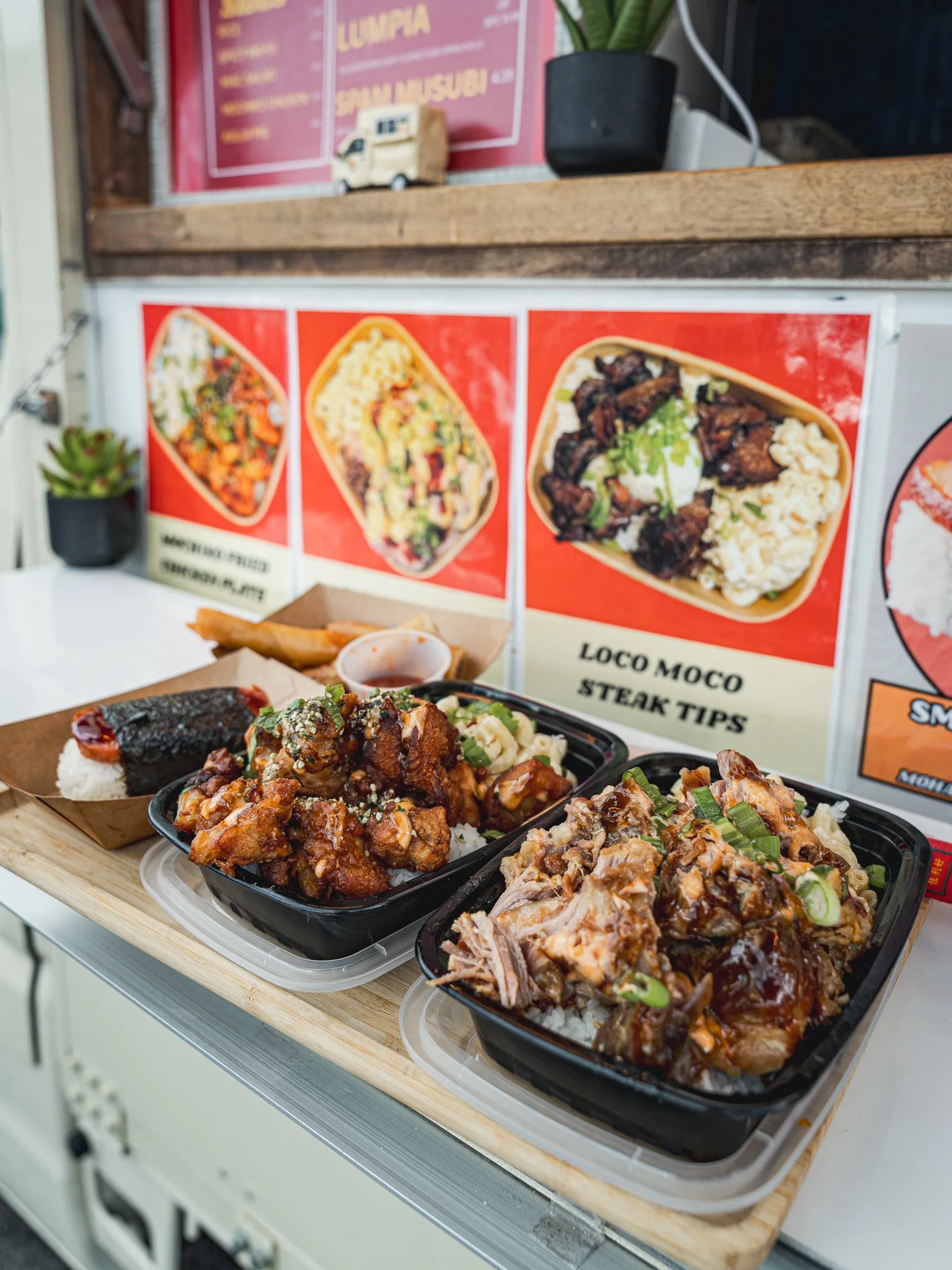 Three black containers filled with Asian-style beef and rice dishes, placed on a wooden tray at a food stall. In the background, a menu with images of similar dishes and a small succulent plant are visible.