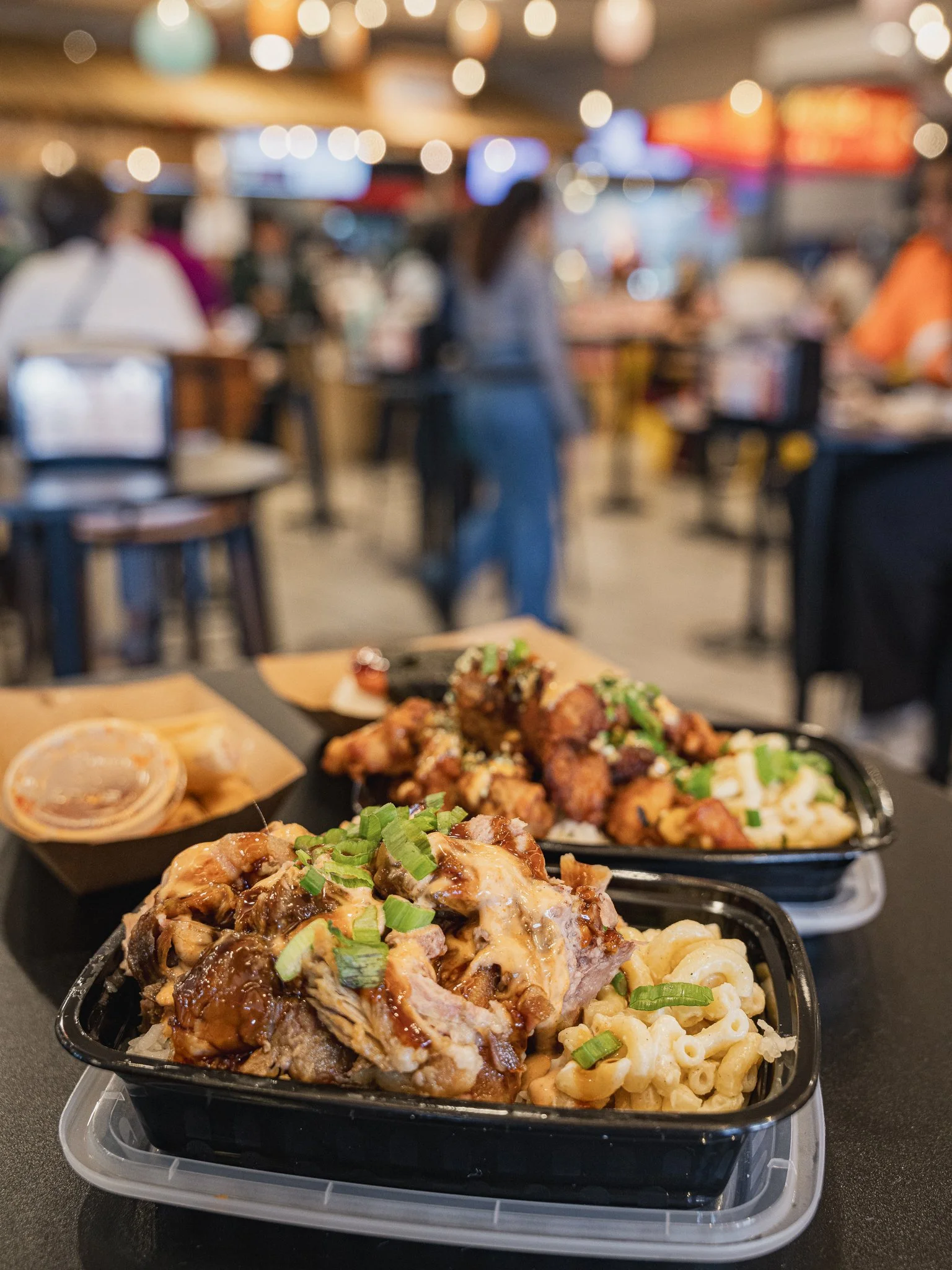 Close-up of takeout trays filled with Asian-style dishes such as noodles, fried chicken, and meat with sauce, set on a table in a restaurant with blurred interior and patrons in the background.