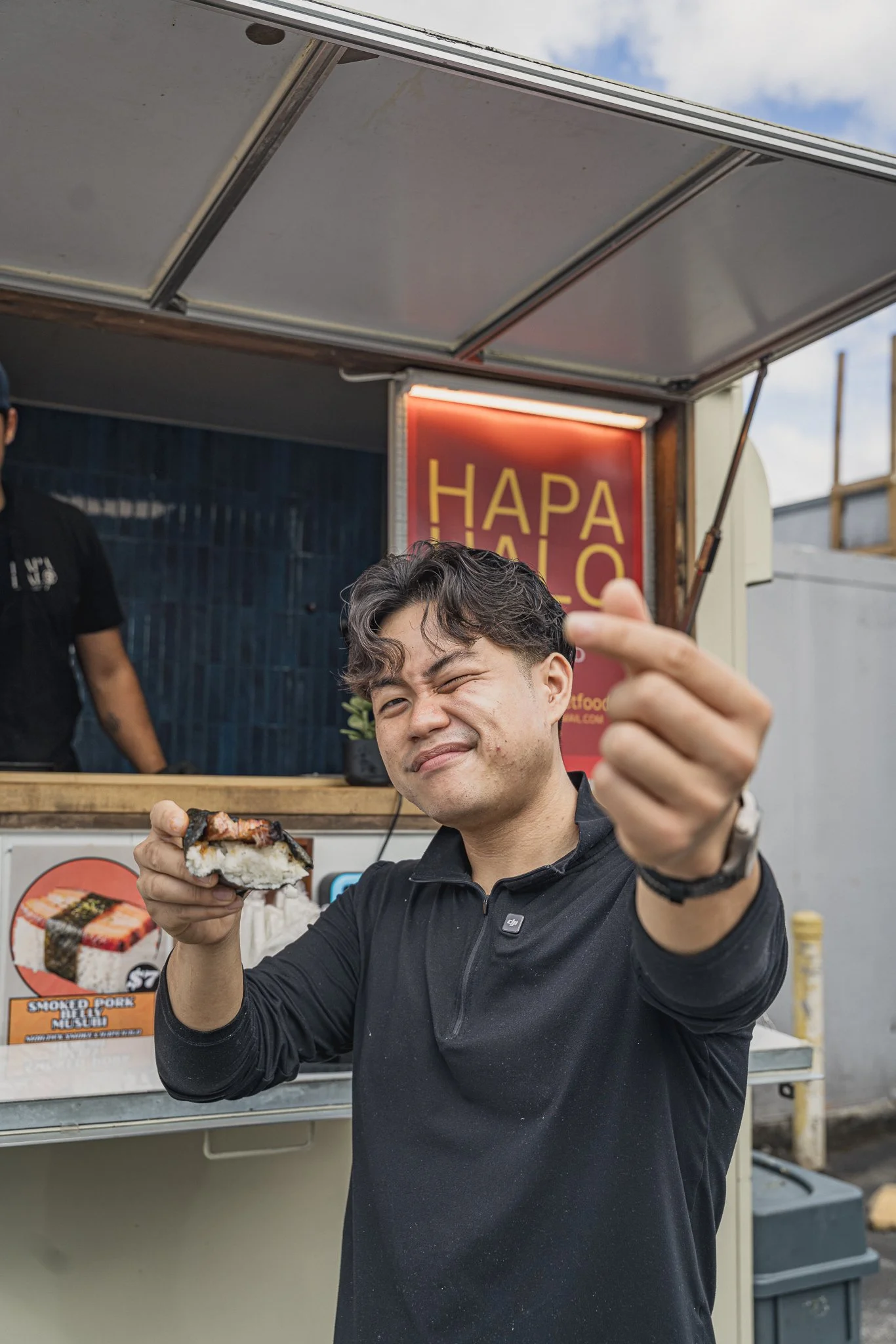 A man with black hair and a black shirt holding a sushi roll and making a peace sign. He is smiling and winking in front of a food stall with a red sign that says "HAPA HAO" and an image of smoked pork musubi.