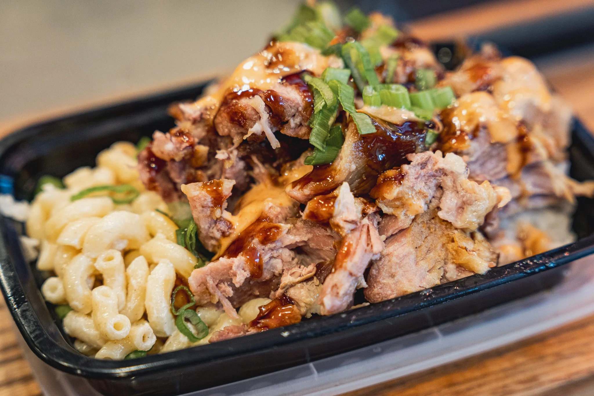 A black takeout container filled with macaroni and cheese on one side and shredded barbecue pork topped with chopped green onions and drizzled with barbecue sauce on the other side.