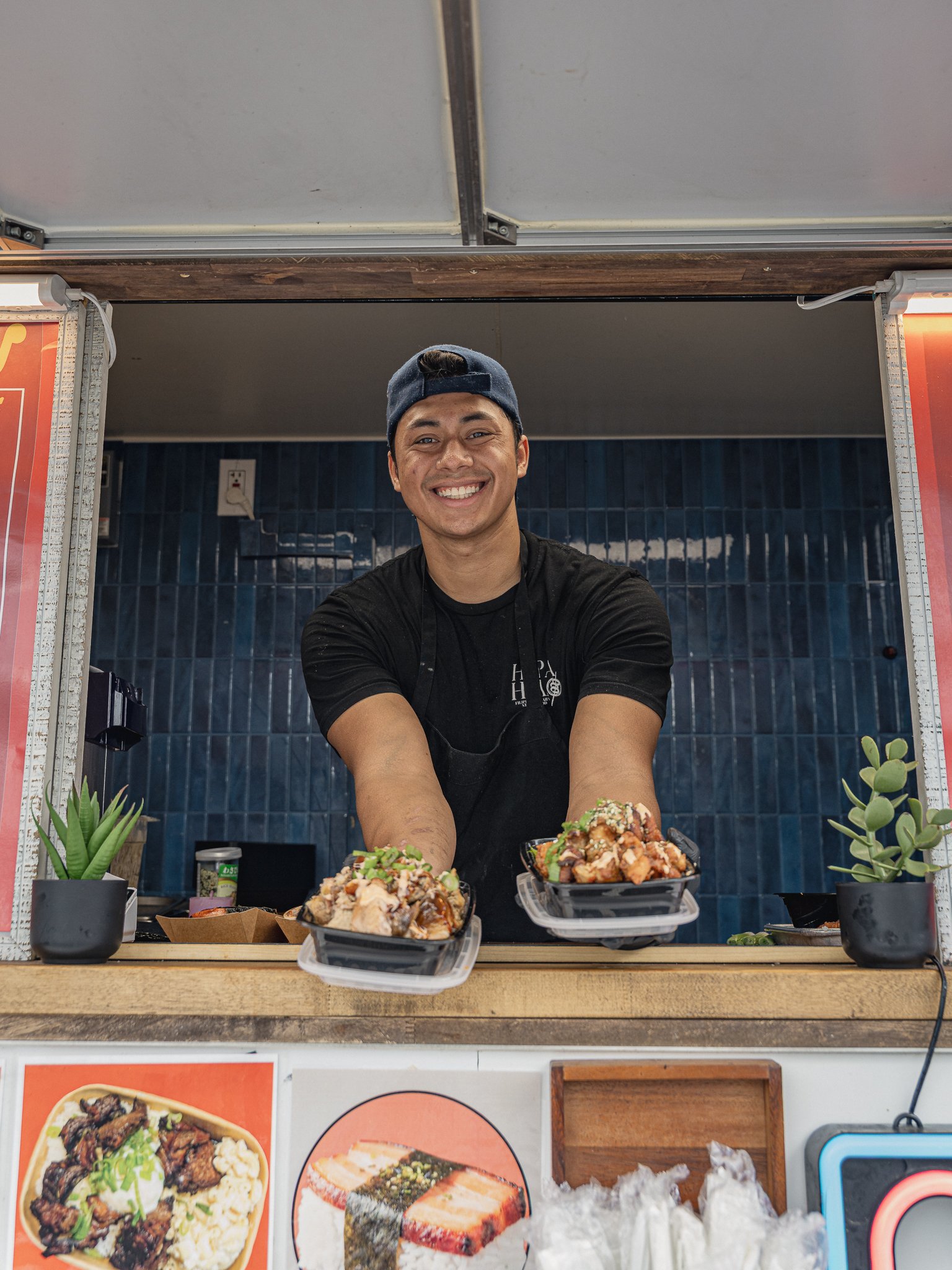 A smiling man wearing a dark cap and black shirt standing behind a food stall with two bowls of food in front of him, surrounded by potted plants and a menu with images of dishes.