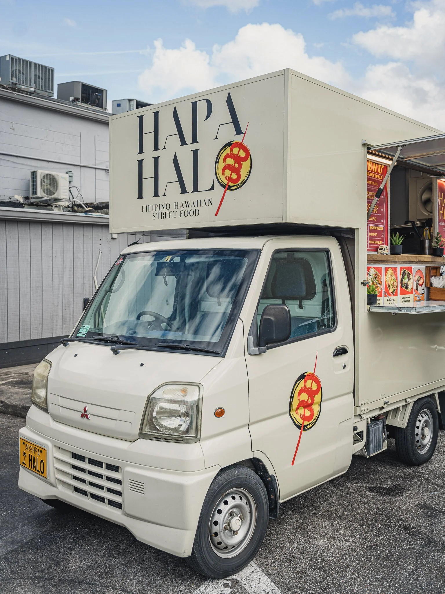 Food truck with a white body and a sign reading 'HAPA HALO' with a pineapple graphic, parked in a lot with a building in the background and a cloudy sky.