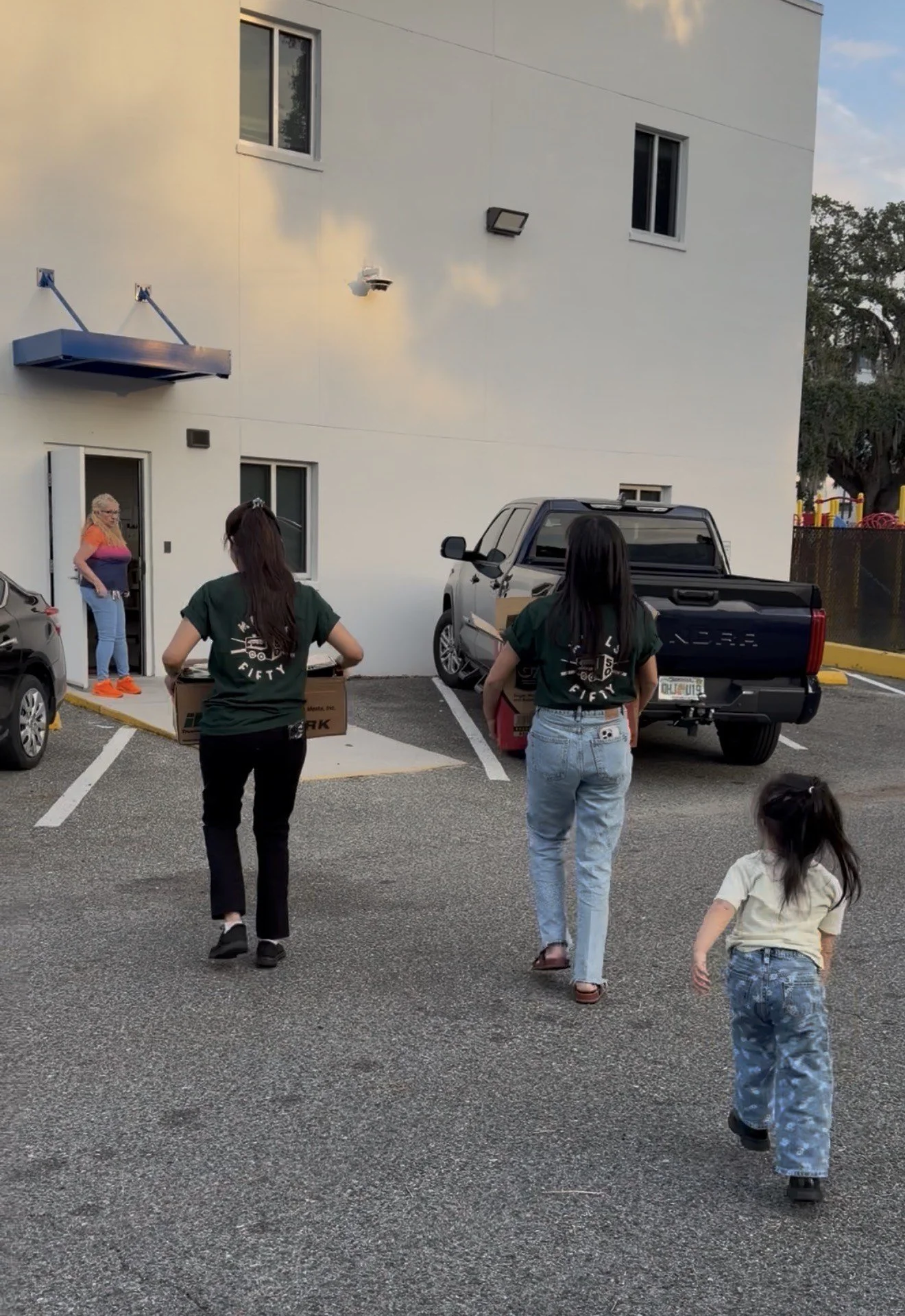 People walking in a parking lot near a white building with two windows. Two women are carrying boxes, and a woman is standing near a door. There is a truck and parked cars, with a playground visible in the background.
