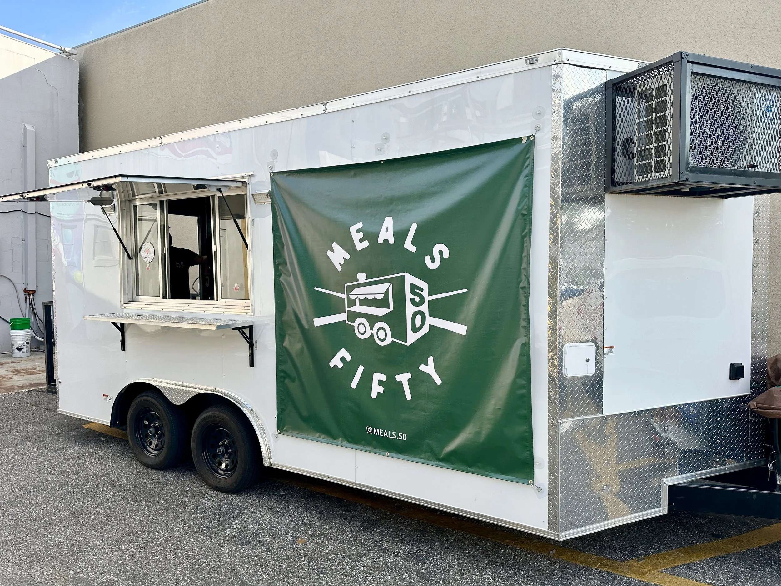 A food truck with a green banner that reads 'Meals 50' and has an illustrated food stand logo. The truck has a window for ordering and a diamond plate metal surface on one side.