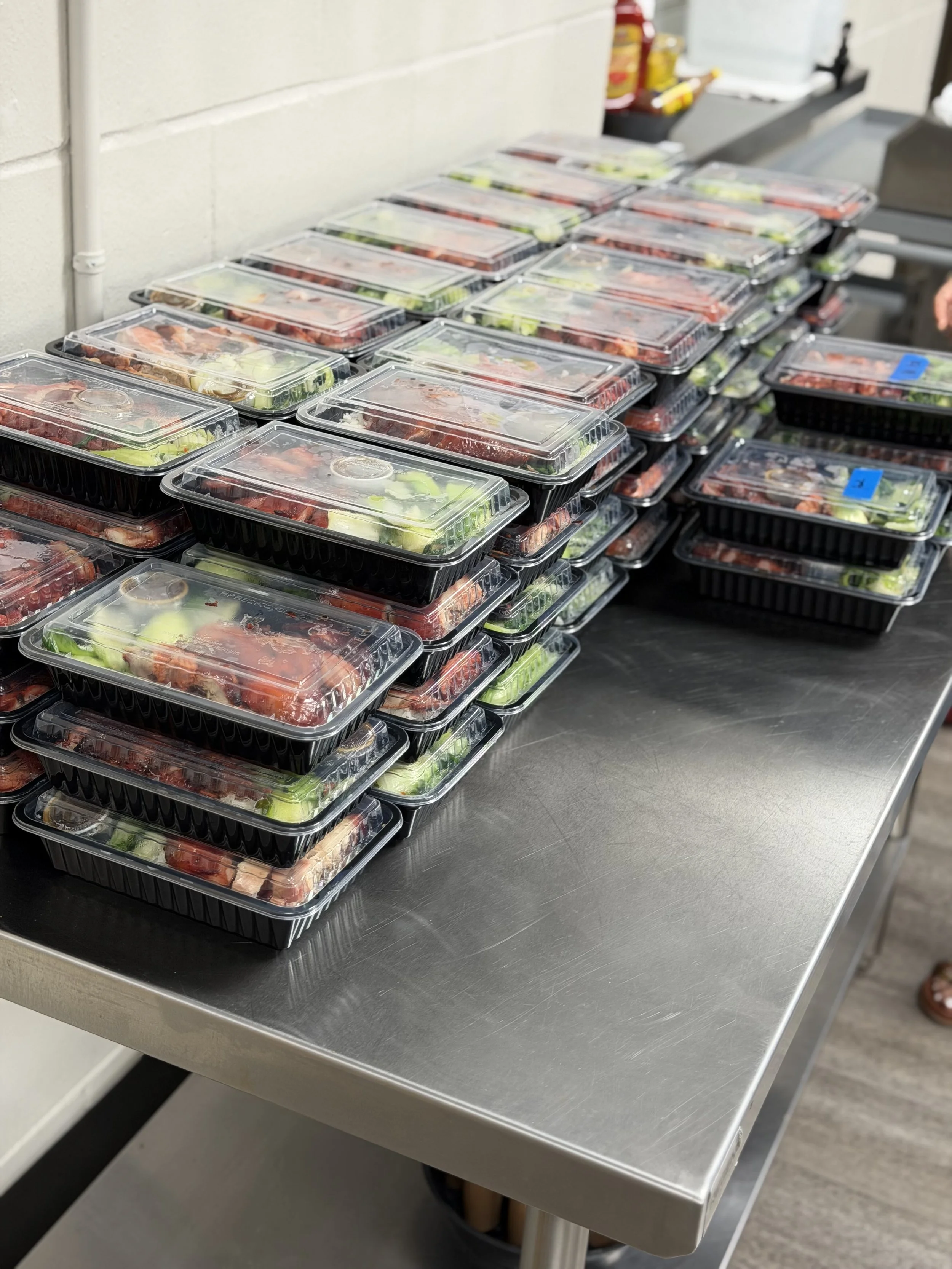 Stacked plastic food containers filled with salads on a stainless steel table at a grocery store or deli.