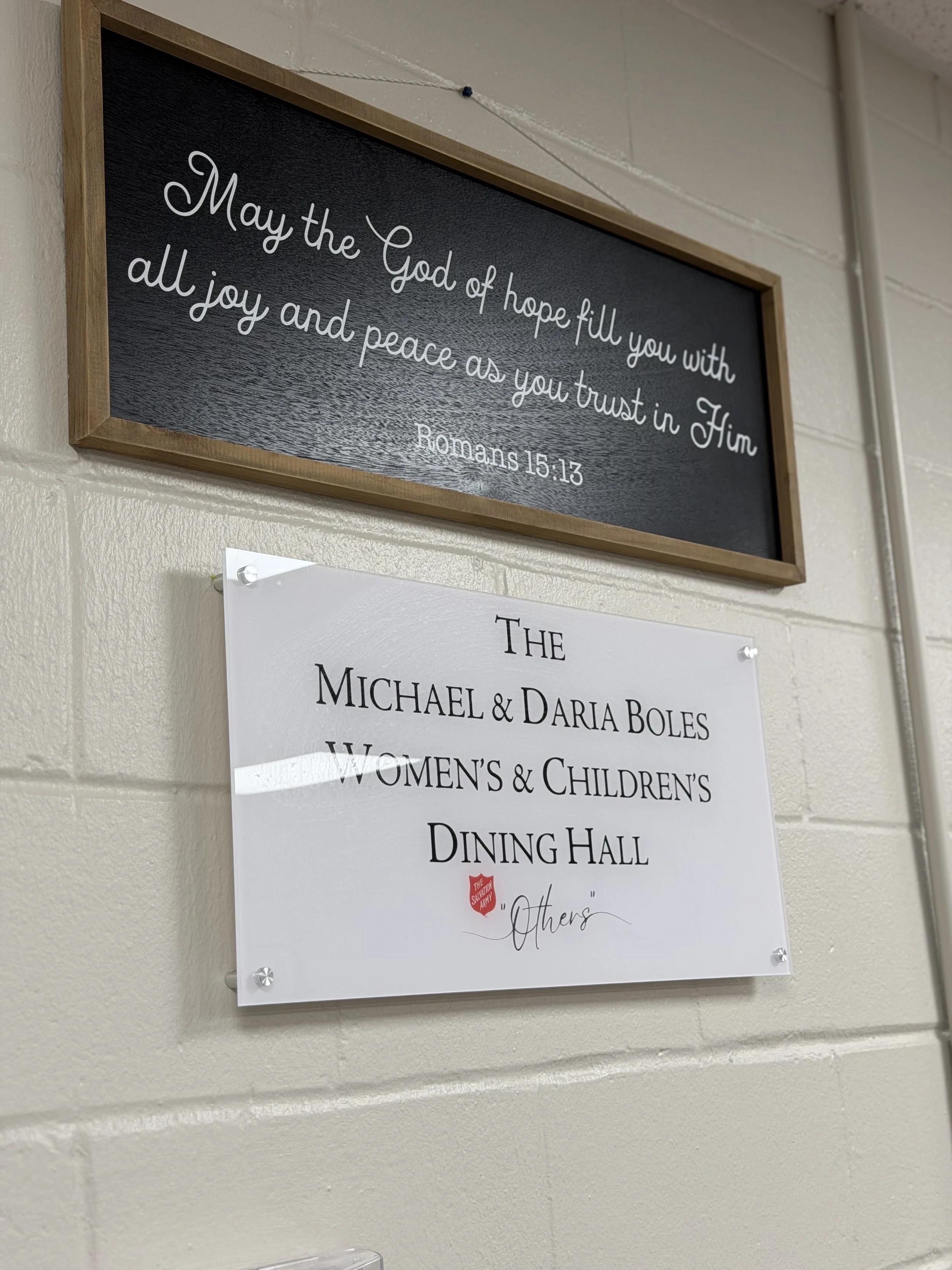 A black framed sign on a beige wall with the quote: "May the God of hope fill you with all joy and peace as you trust in Him Romans 15:13."