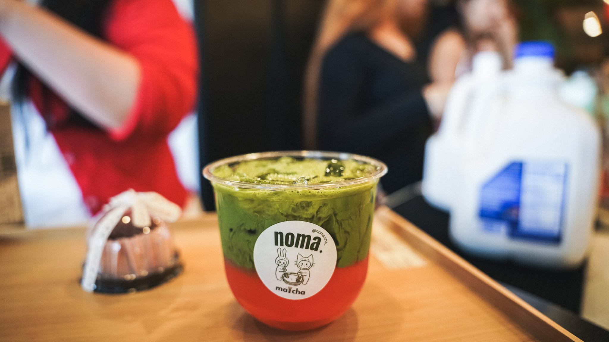 A plastic cup of green matcha latte with ice cubes and a red bottom, labeled "noma. matcha" with cartoon characters, on a wooden table in a cafe setting.