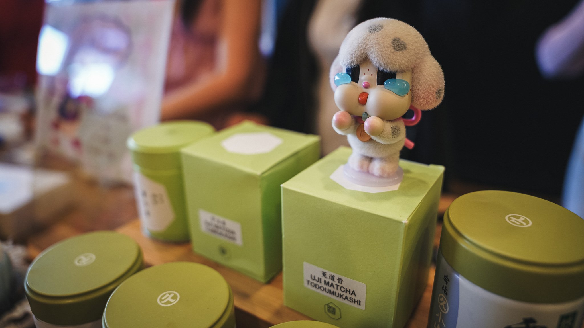 A plush doll with tearful eyes and a sad expression, displayed on a green box at a market or store. Surrounding it are various green and yellow containers, some labeled with Japanese text.