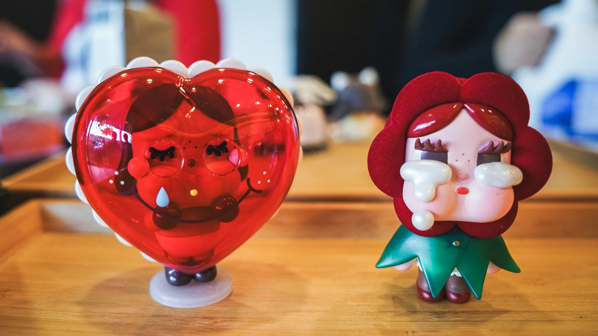 Two decorative figurines on a wooden surface, one resembling a crying heart with a face, and the other a girl with a large red bow on her head, tears on her face, and a green dress.