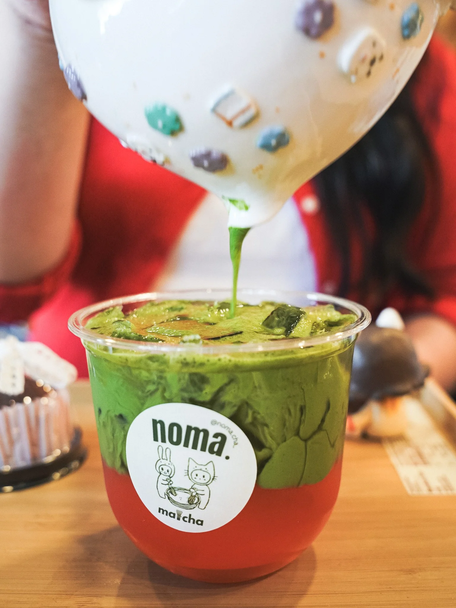 A clear plastic cup filled with a green beverage, likely matcha, with ice cubes inside. The cup has a white circular logo featuring cartoon animals and the word 'noma'.