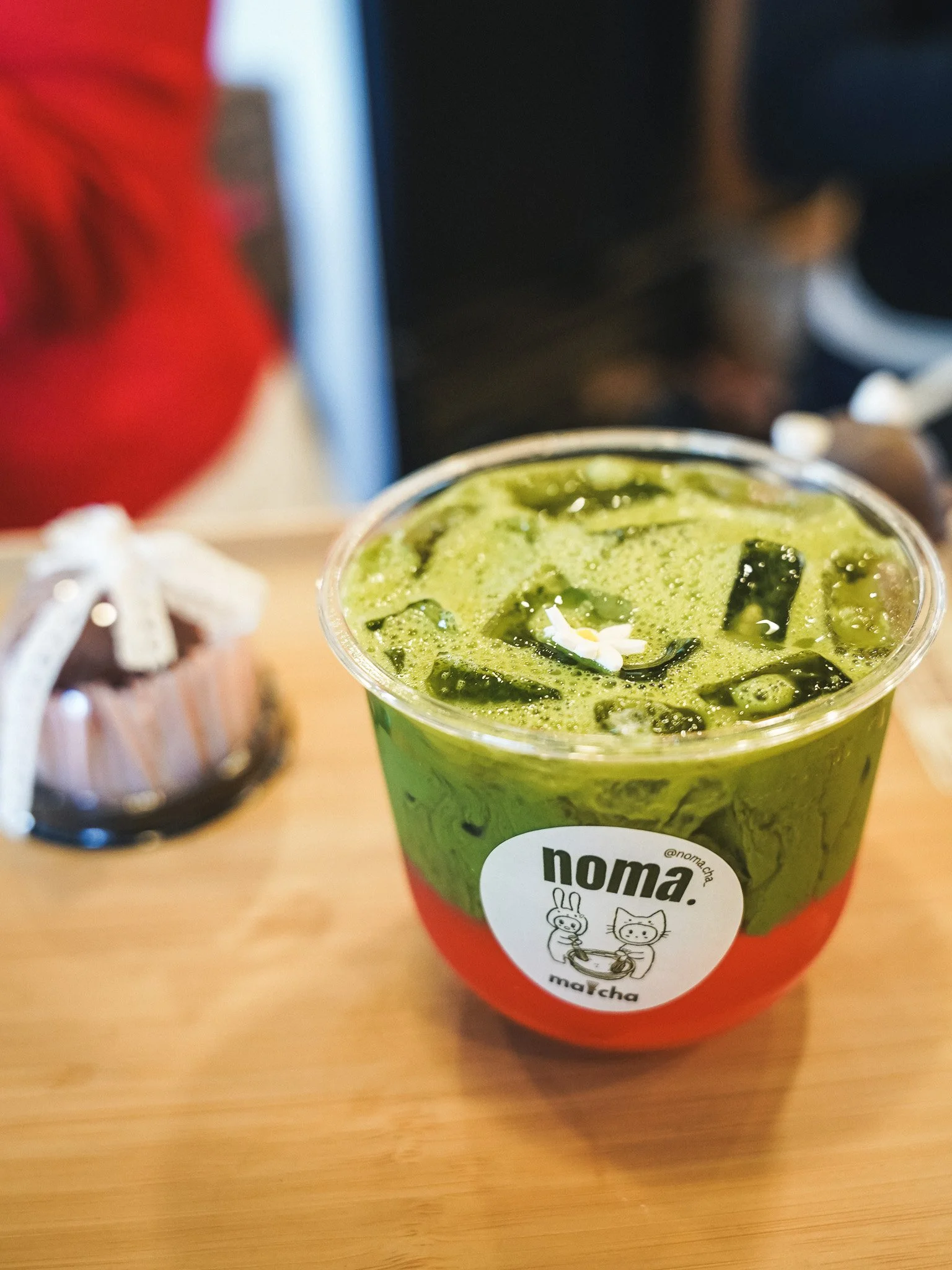 A clear plastic cup filled with a green matcha-flavored beverage topped with dark seaweed strips and a small white flower, placed on a wooden table, with a blurred background.