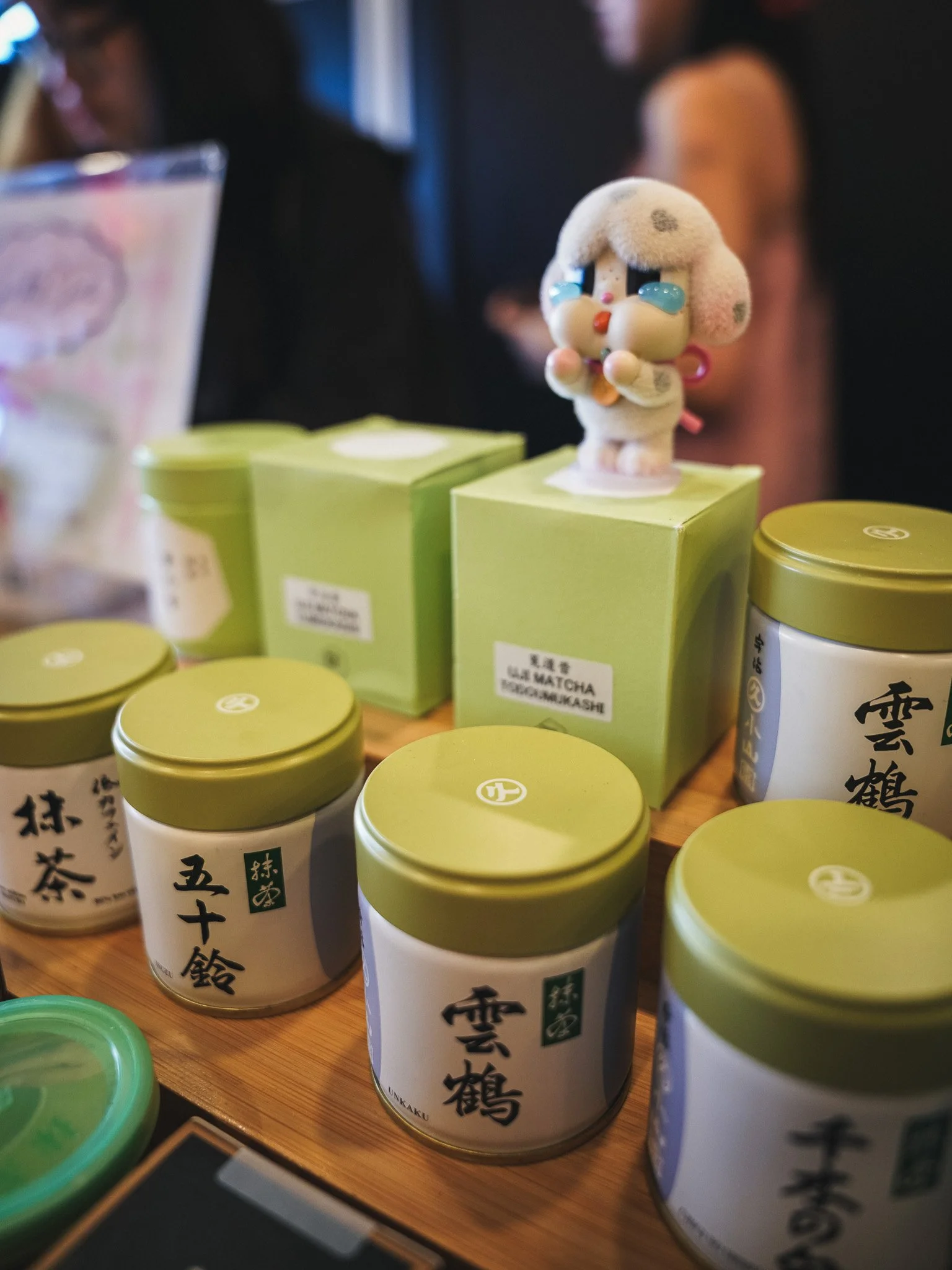 Close-up of several white cylindrical containers with green lids, displaying Japanese characters, on a wooden surface at a store or market. A plush toy with a cute face, blue tears, and pink accessories stands on top of one container.