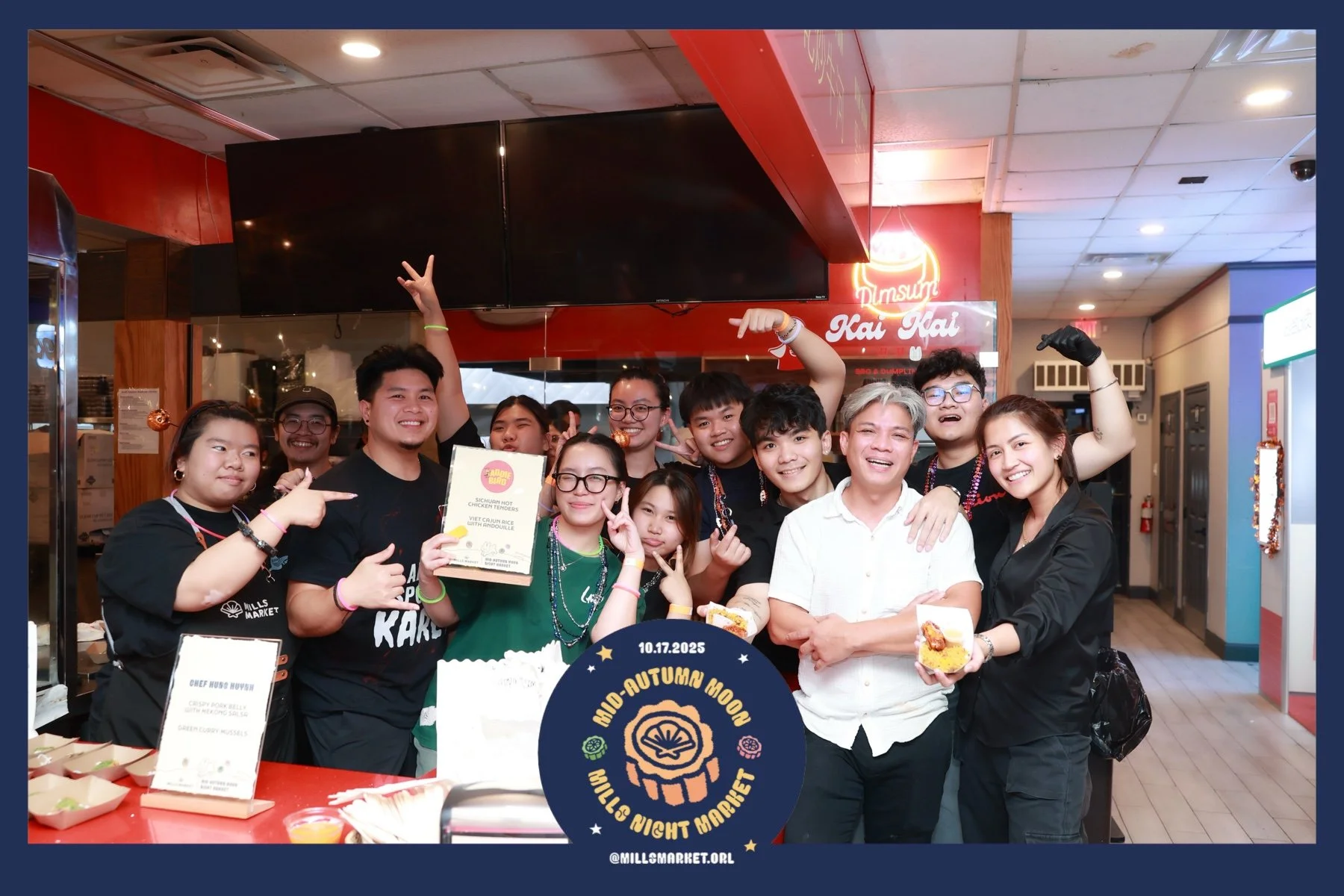 A group of people at a restaurant celebration, smiling and posing for a photo, with some holding food and a certificate, in front of a sign that reads 'Mills Market Autumn Moon.'