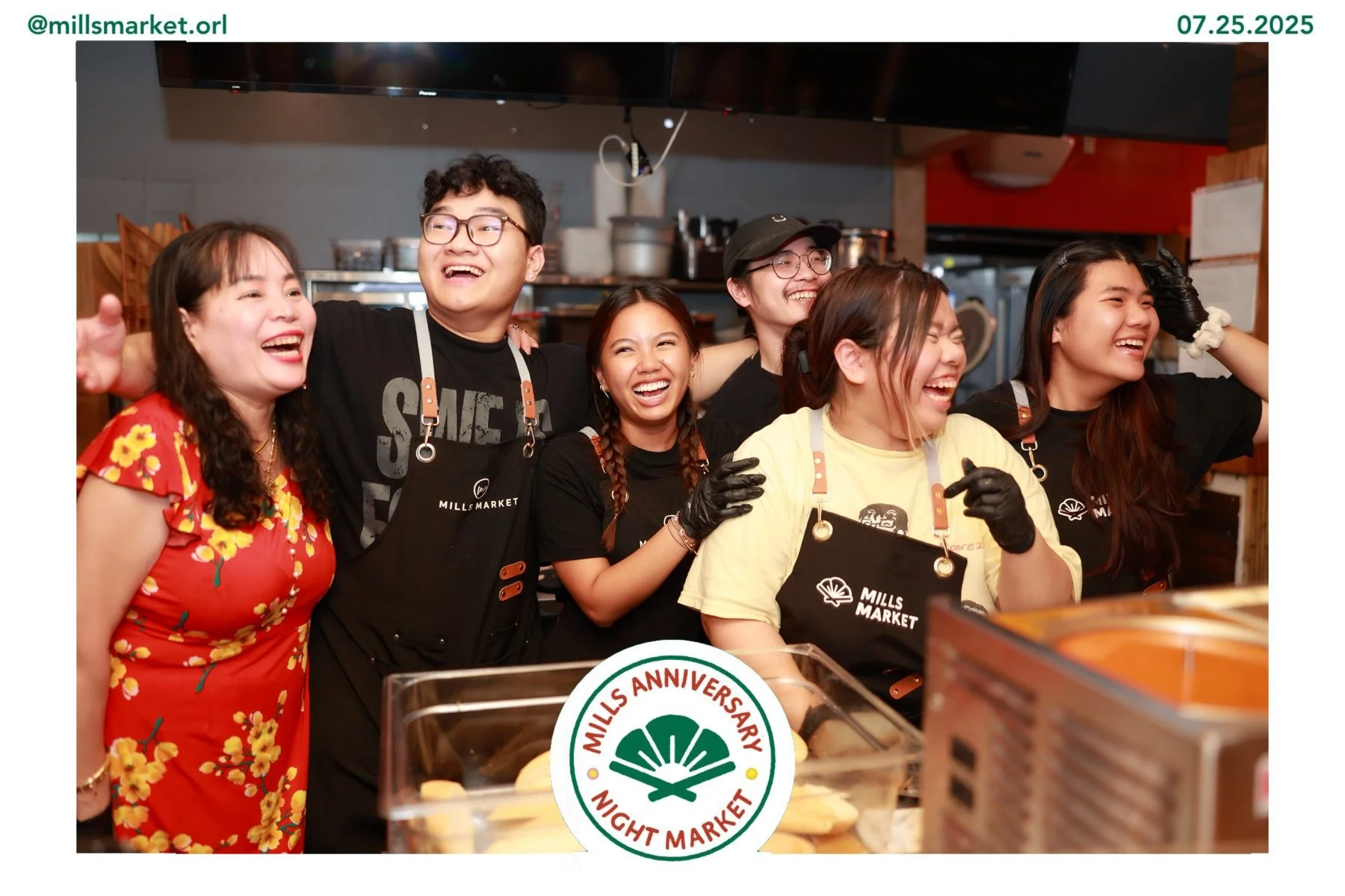 Group of six smiling young adults in a kitchen celebrating Mills Market's anniversary, wearing aprons and casual clothing.
