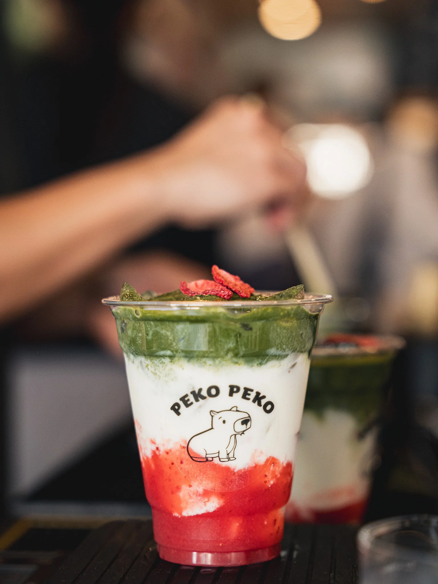 A colorful layered drink in a clear cup with a cartoon dog logo and the words 'PEKO PEKO', topped with crushed red and green toppings.