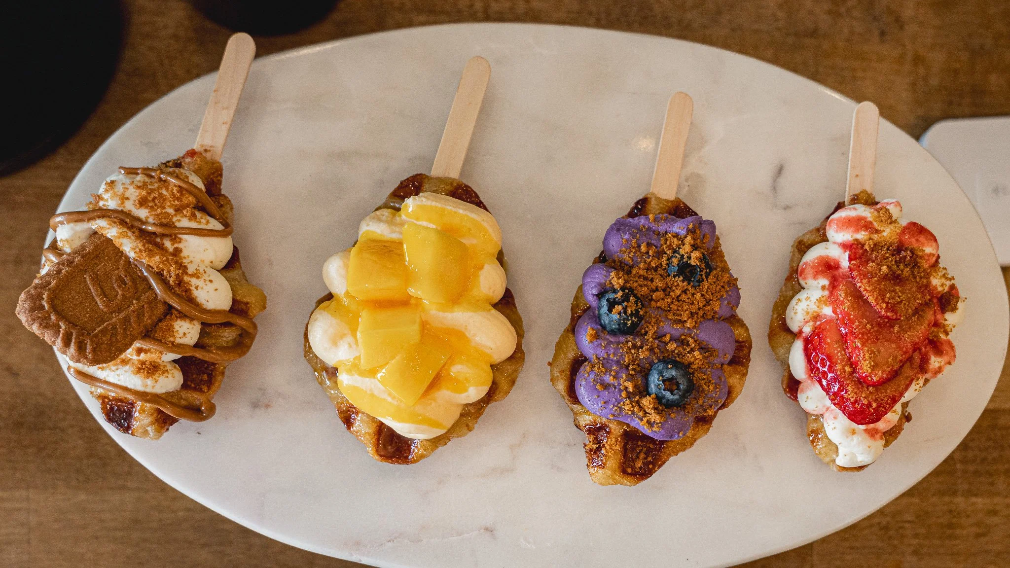 Four pieces of ice cream waffle frites topped with different toppings, arranged on a white oval plate.