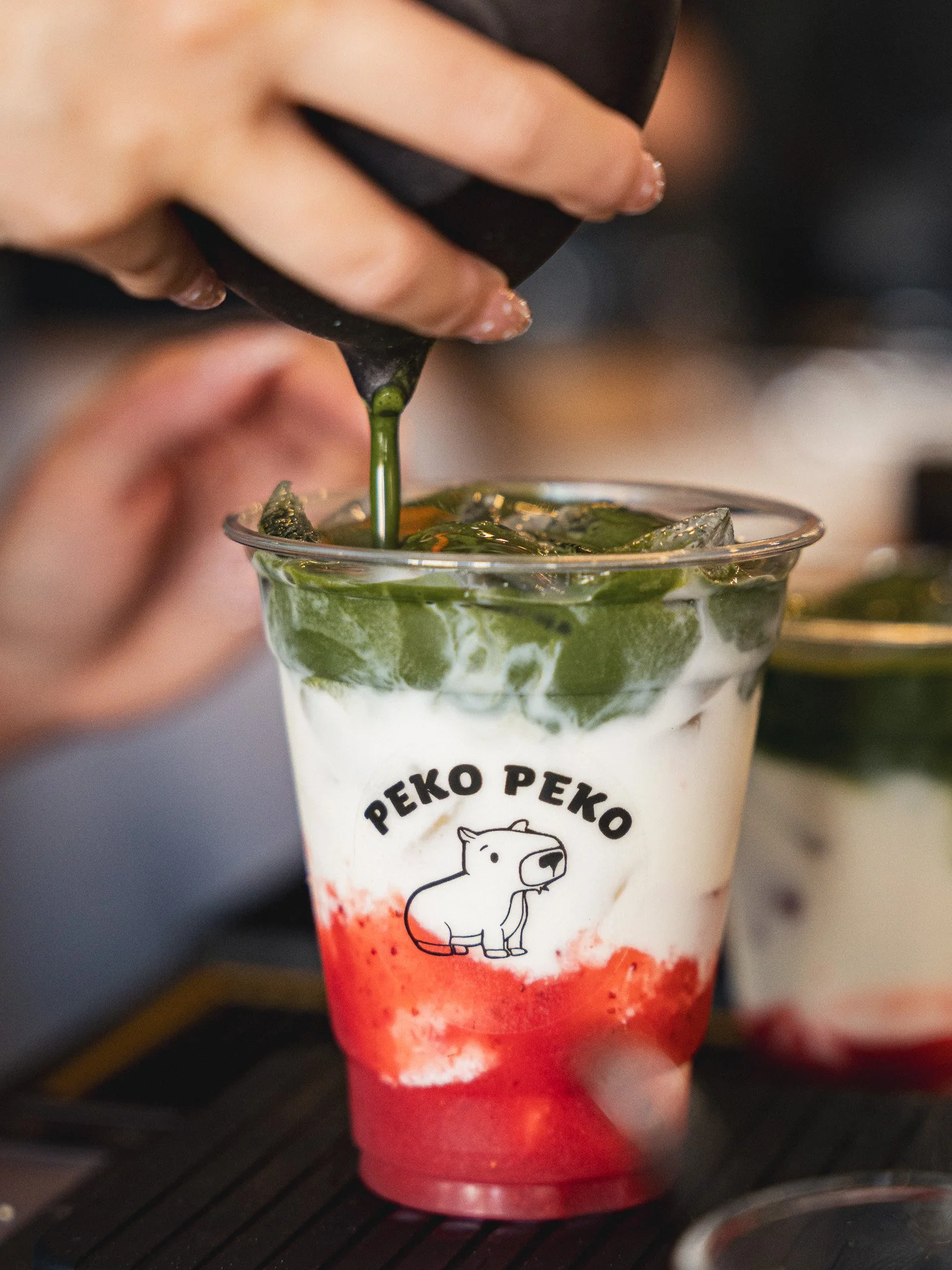 A plastic cup of layered beverage with green matcha, white cream, and red syrup, featuring a cartoon dog logo and Japanese text.