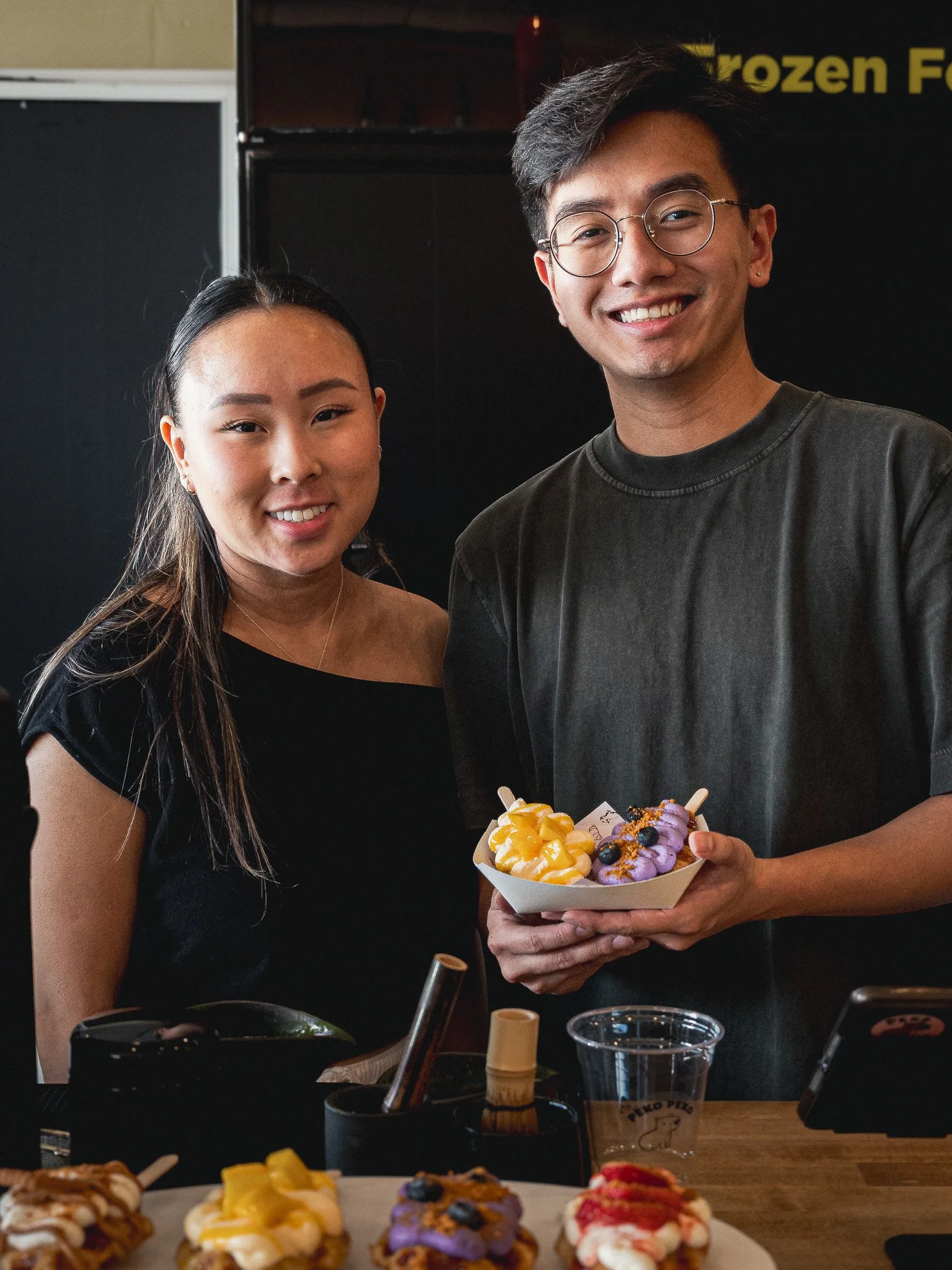 Two people smiling at a table with assorted colorful desserts, with one person holding a tray of purple, yellow, and mixed fruit-topped donuts.
