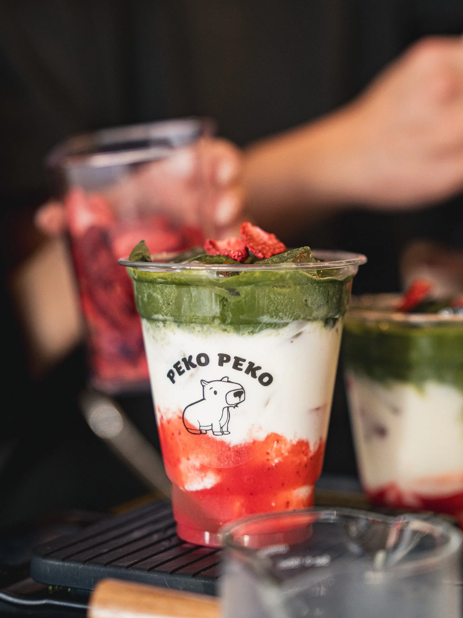 A dessert drink in a clear glass with a cartoon bear logo that reads 'PEKO PEKO'. The drink has a green and red layered appearance, topped with strawberries and a green topping, possibly matcha or spinach powder. The glass is placed on a dark surface