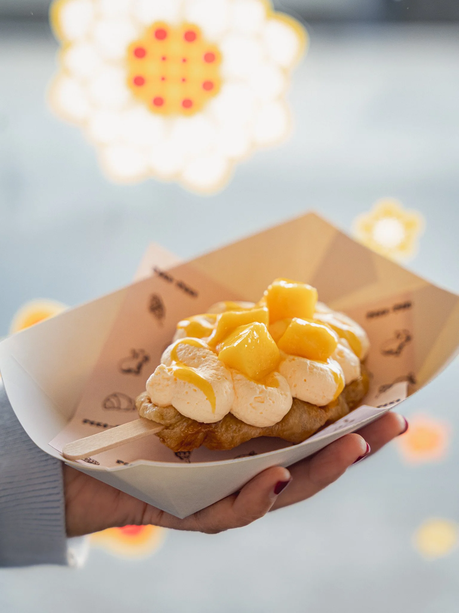 Close-up of a hand holding a dessert topped with fresh mango chunks and a drizzle of yellow sauce, served in a paper tray with a wooden stick.