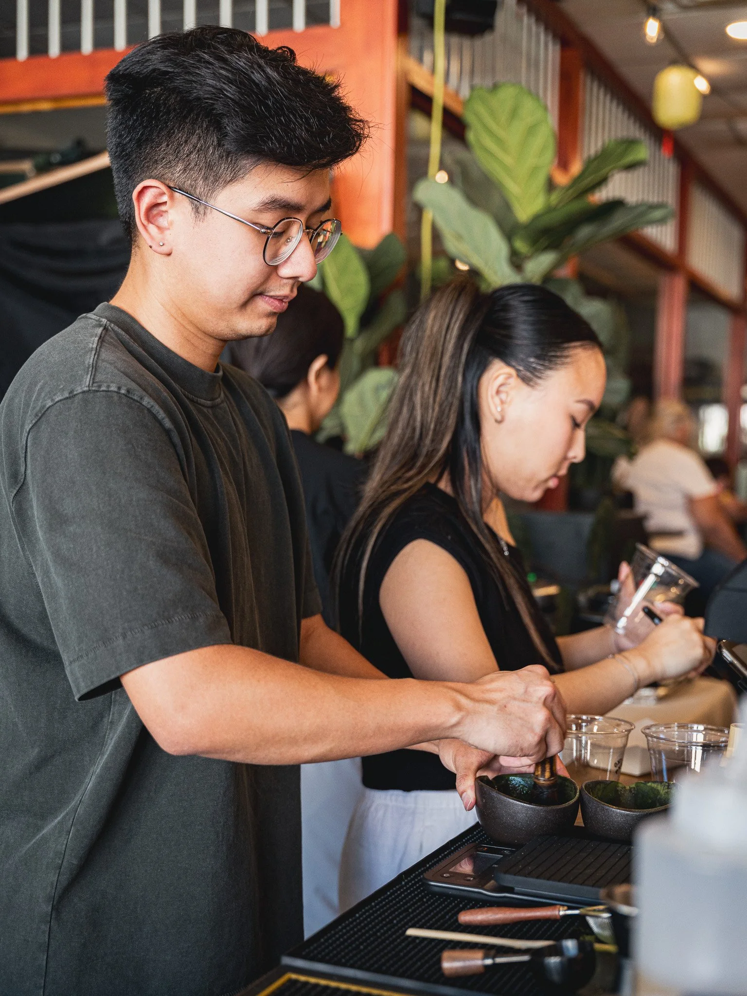 A man with black hair, glasses, and earrings is preparing food at a counter, holding a bowl and spoon. A woman with long dark hair is next to him, looking at her phone. The counter has various kitchen utensils and bowls, with a background of large gr