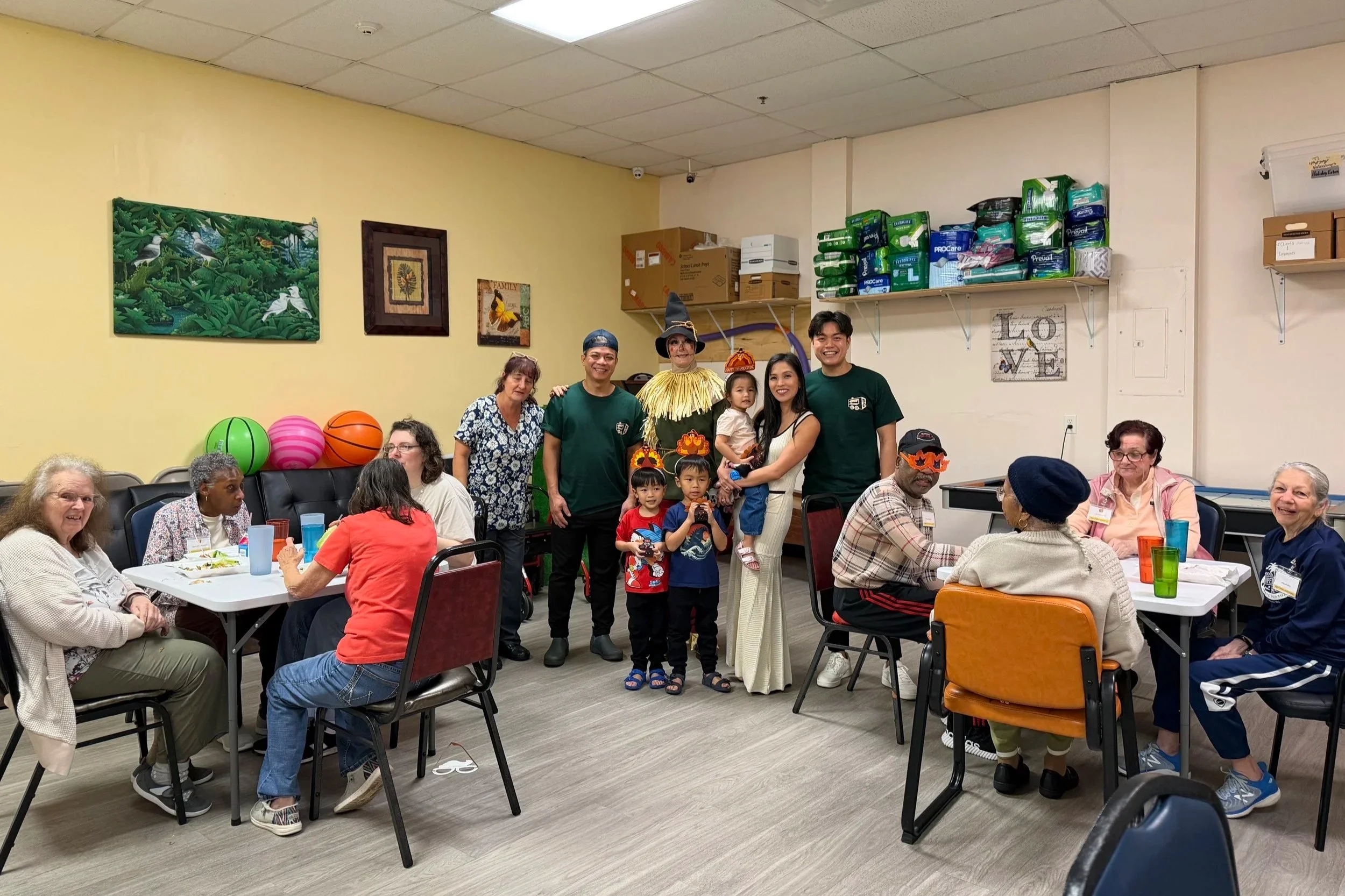Group of diverse people, including children and adults, gathered in a community room for a celebration. Some are wearing costumes, and everyone is smiling. The room is decorated, with colorful balls on a table and framed artwork on the wall.