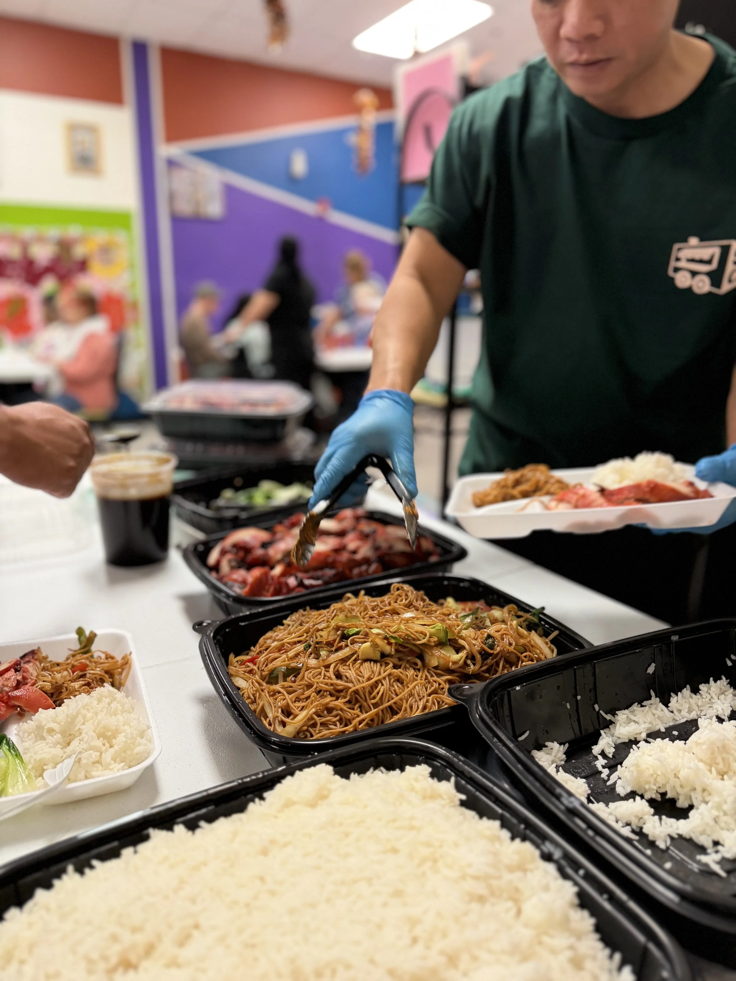 A person serving Asian cuisine at a buffet, with various dishes including fried rice, noodles, and vegetables in black containers on a table.
