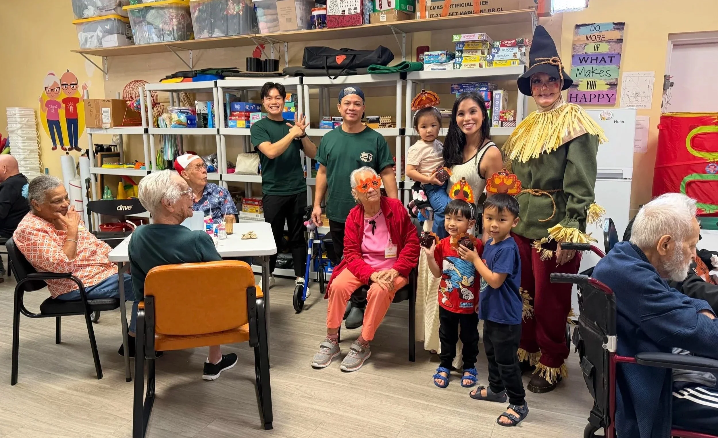 Group of elderly residents and children gathered in a room with some people in costume. They are surrounded by shelves filled with toys and supplies. Some residents are seated, chatting, while others are standing and smiling for the camera, which cap