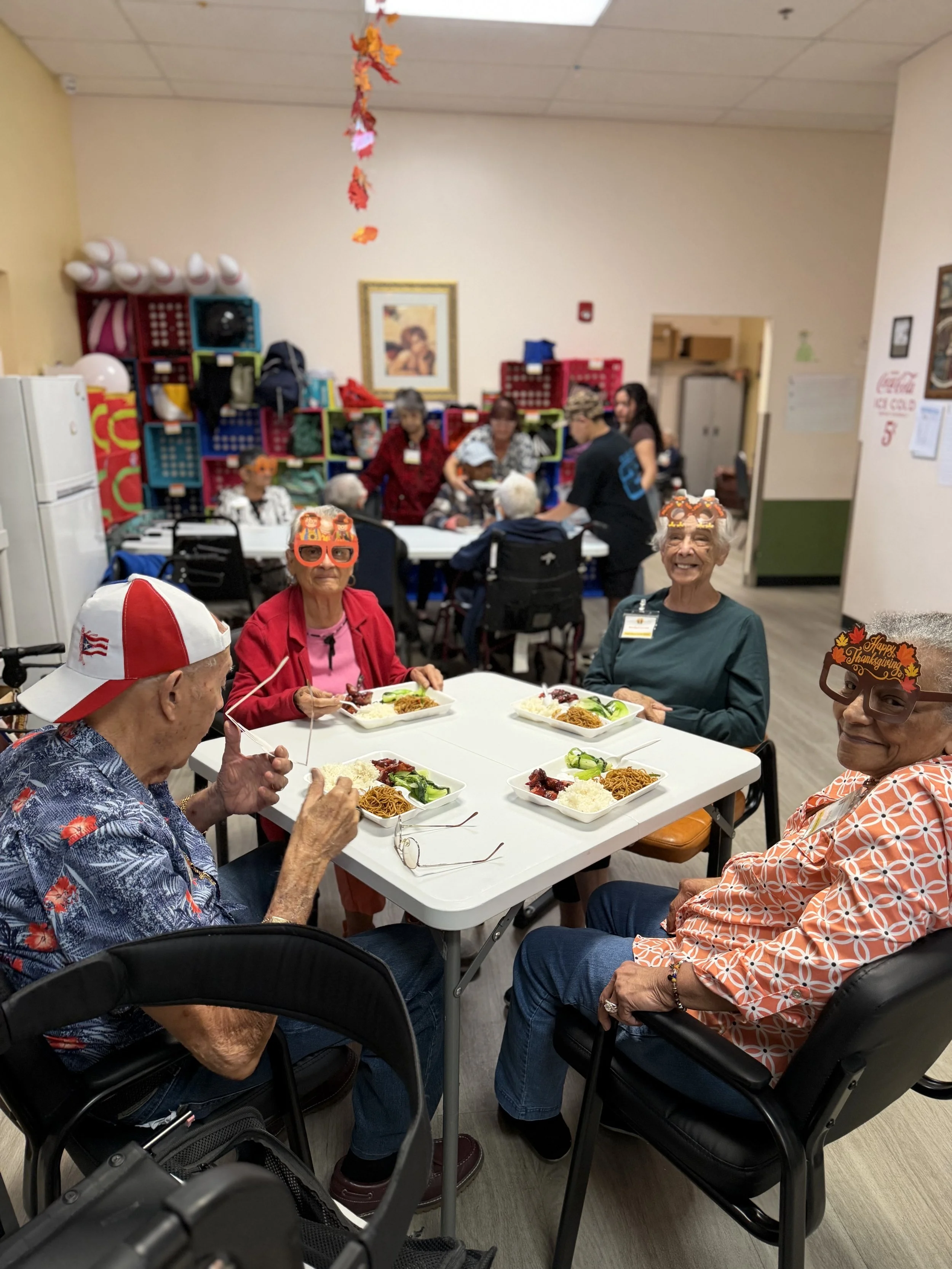 Elderly people at a Thanksgiving celebration, sitting at a table with plates of food, wearing festive glasses and hats, in a community room decorated for fall.