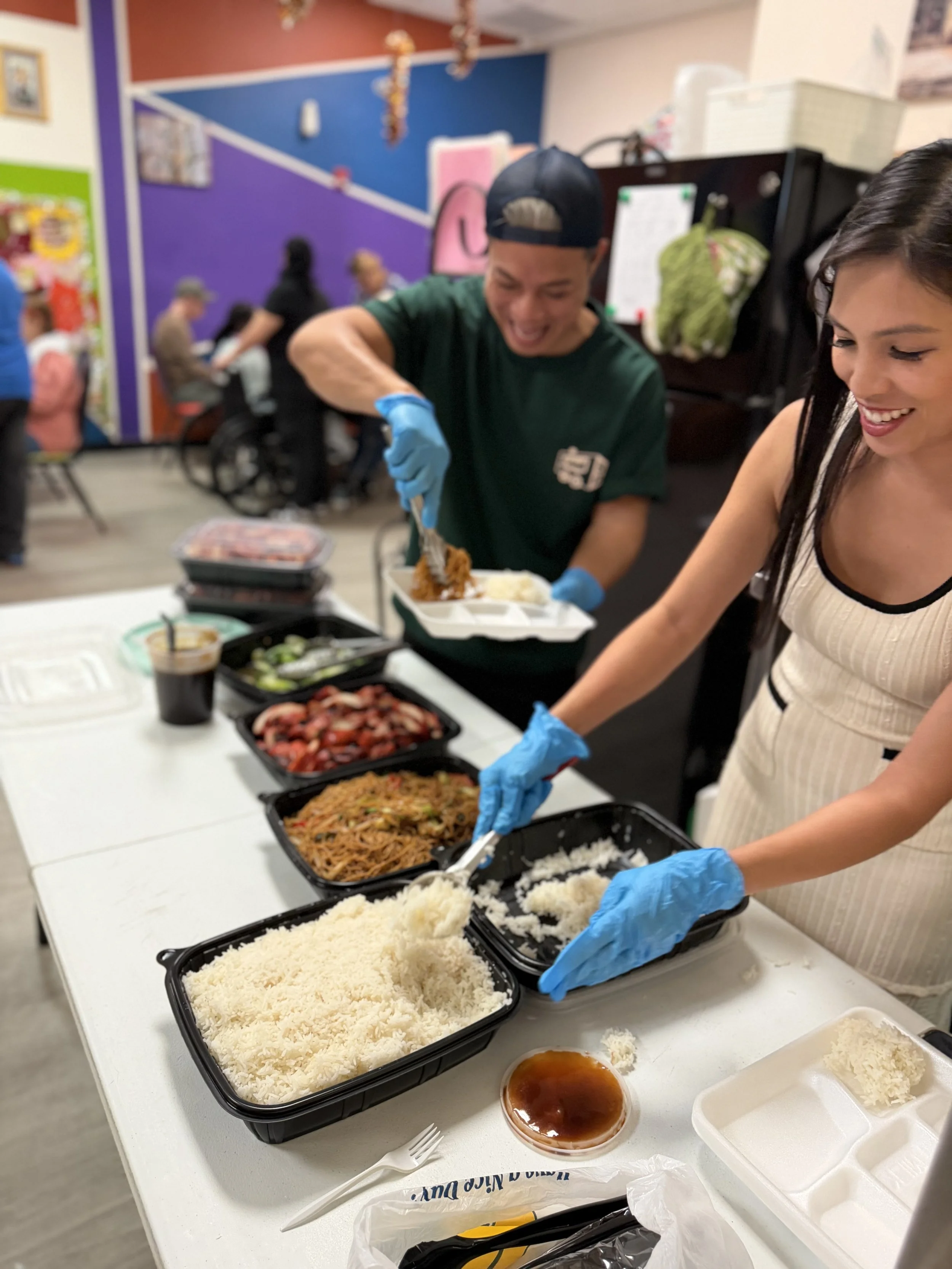 People serving and preparing food in a cafeteria-style setting, with takeout containers and various dishes on the table.