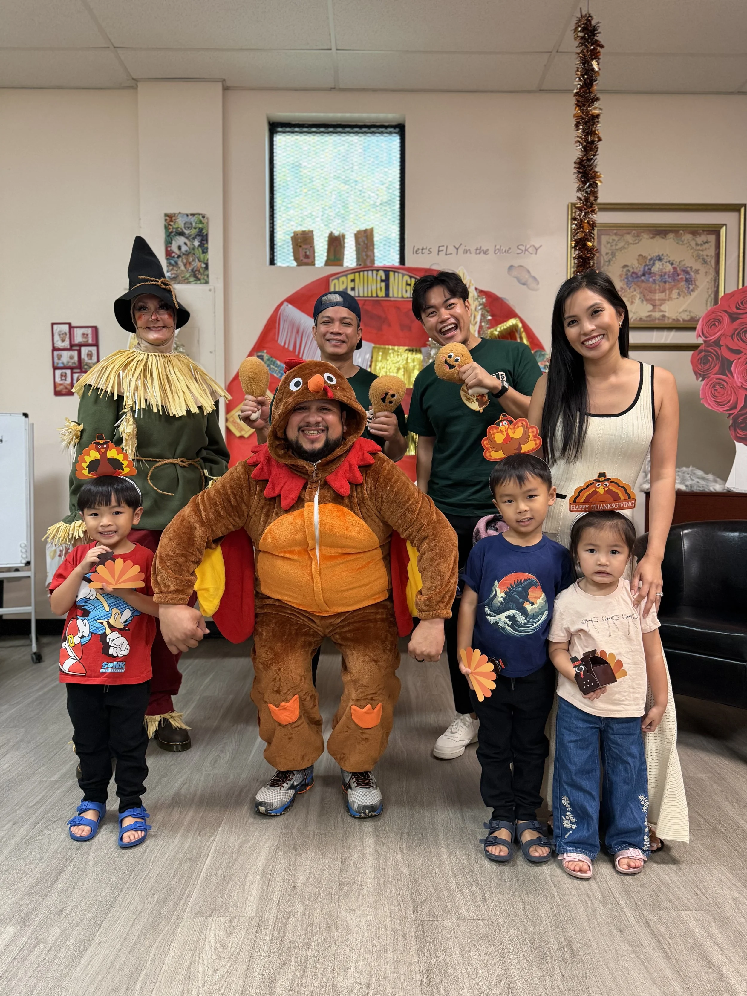 Group of people at a celebration with a person in a turkey costume, children, and adults, some holding Thanksgiving-themed props, in a decorated room.