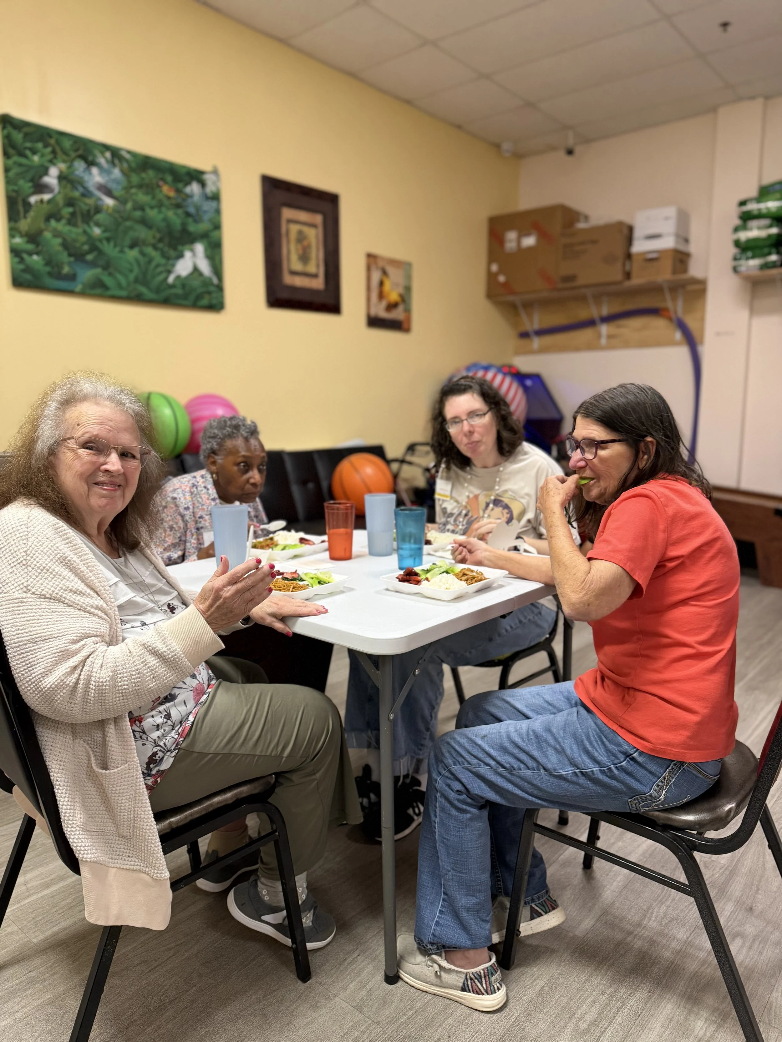 Four women sitting around a table eating a meal in a room with artwork on the wall and storage shelves in the background.