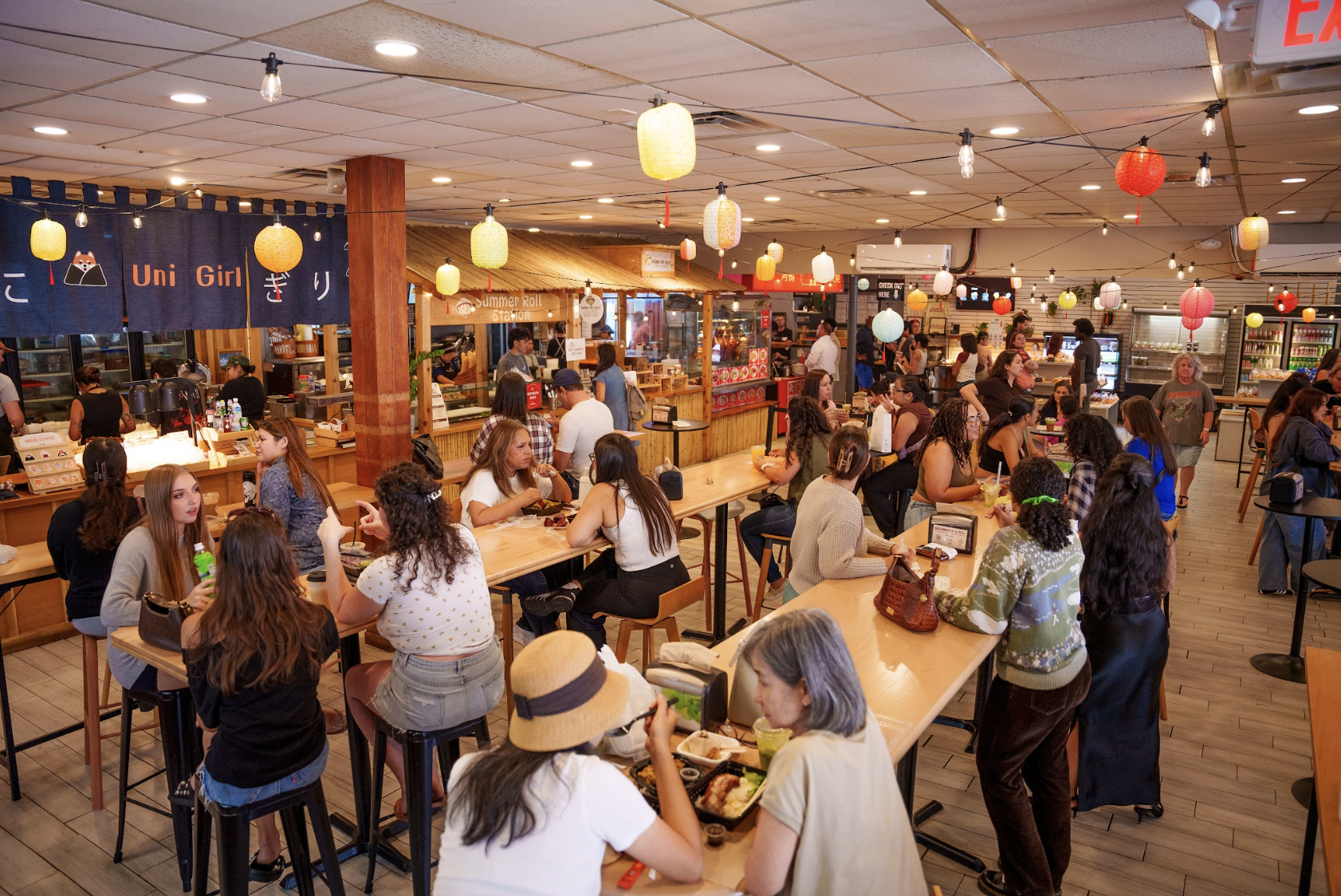A busy indoor food court with many people sitting at tables and eating, festive paper lanterns and string lights hanging from the ceiling, and various food vendors in the background.