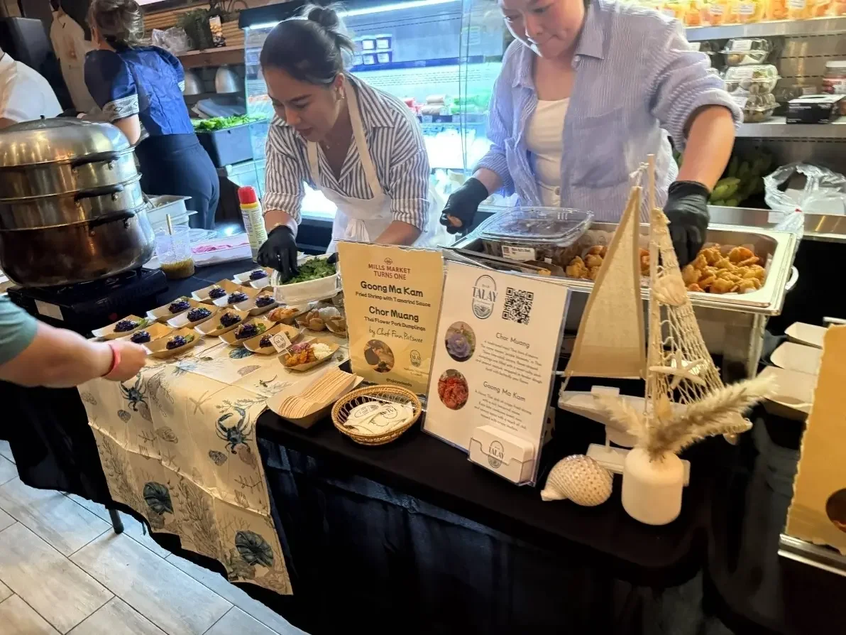 Two women preparing and serving traditional Asian dishes at a market stall, with small bowls of food and informational signs on the table.