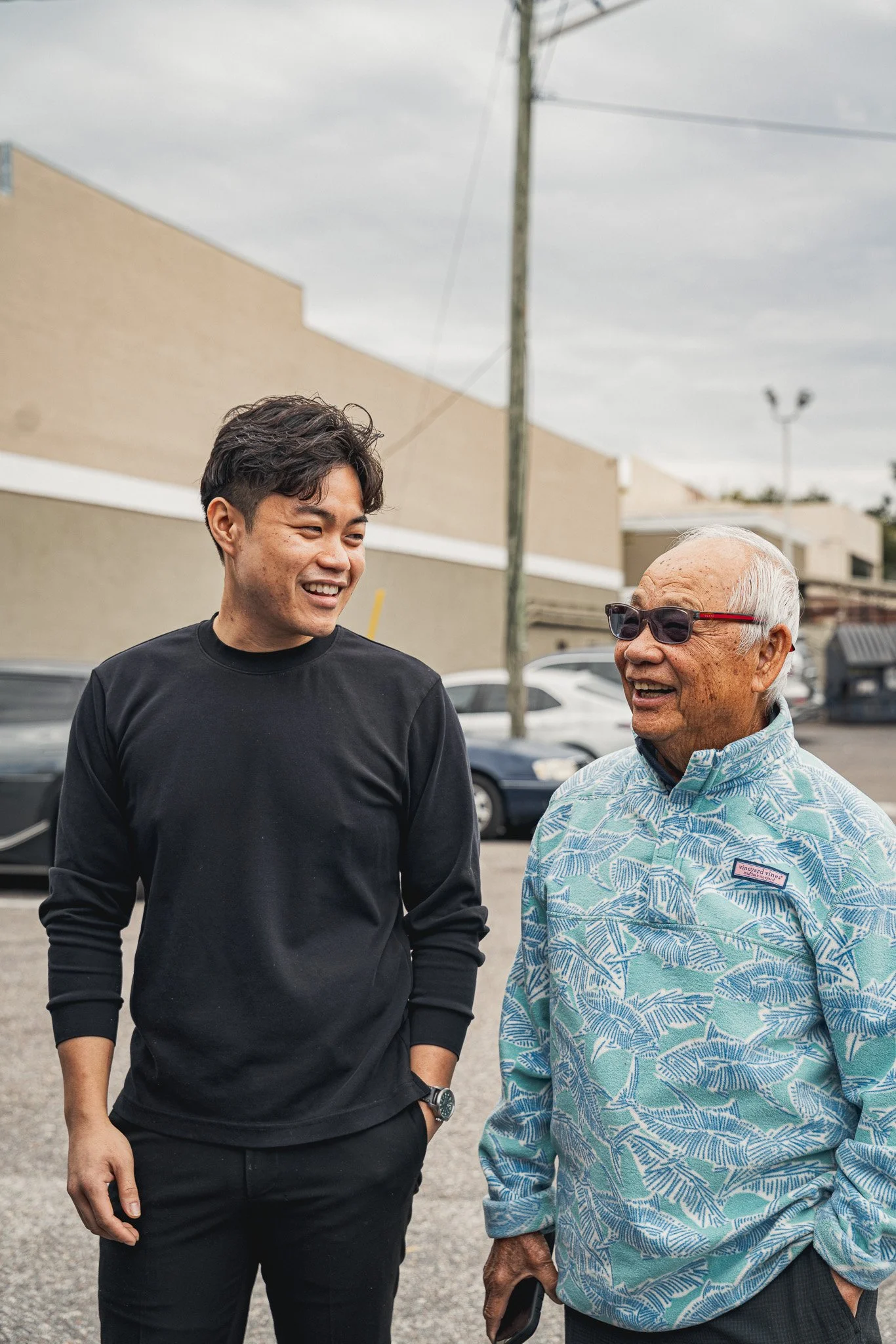 Two men are talking outdoors in a parking lot, smiling at each other. The younger man has short dark hair and is wearing a black long-sleeve shirt. The older man has gray hair, is wearing sunglasses, and a light blue, green, and white patterned butto